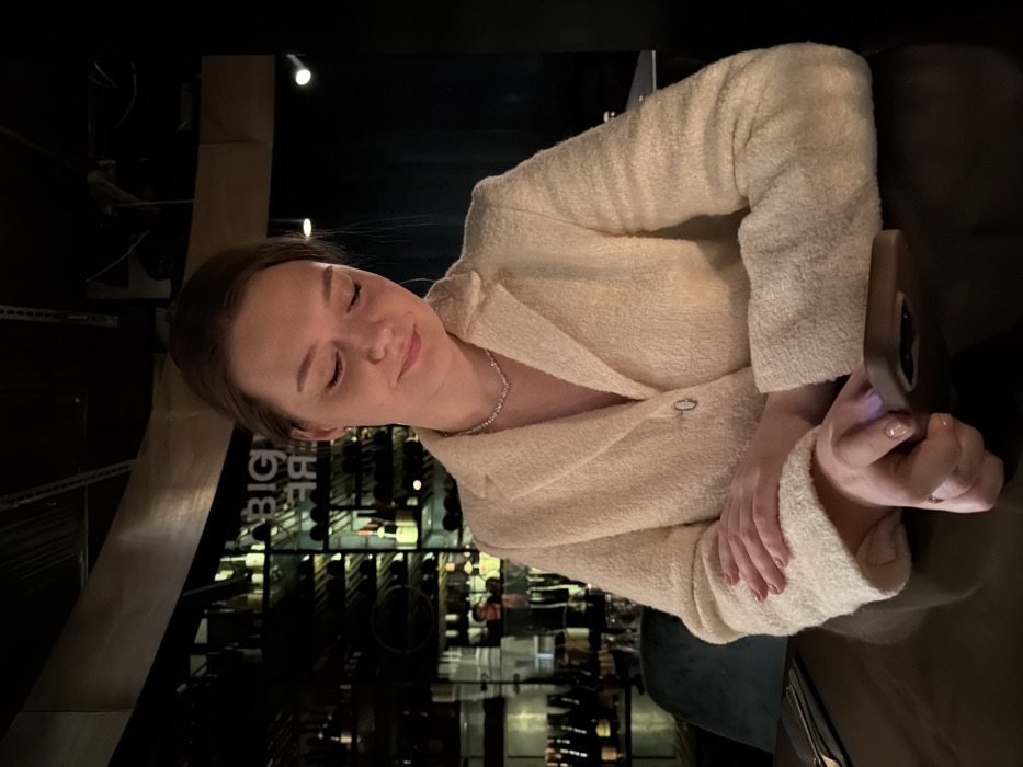 A woman in a cream-colored jacket sits at a table, eyes closed with a serene expression. She holds a phone in her hands. The background is dark, with wine racks visible. The environment suggests a restaurant or bar with a wine cellar. The jacket has a textured, possibly bouclé, fabric. The woman wears a delicate necklace. The lighting is dim and creates a relaxed mood.