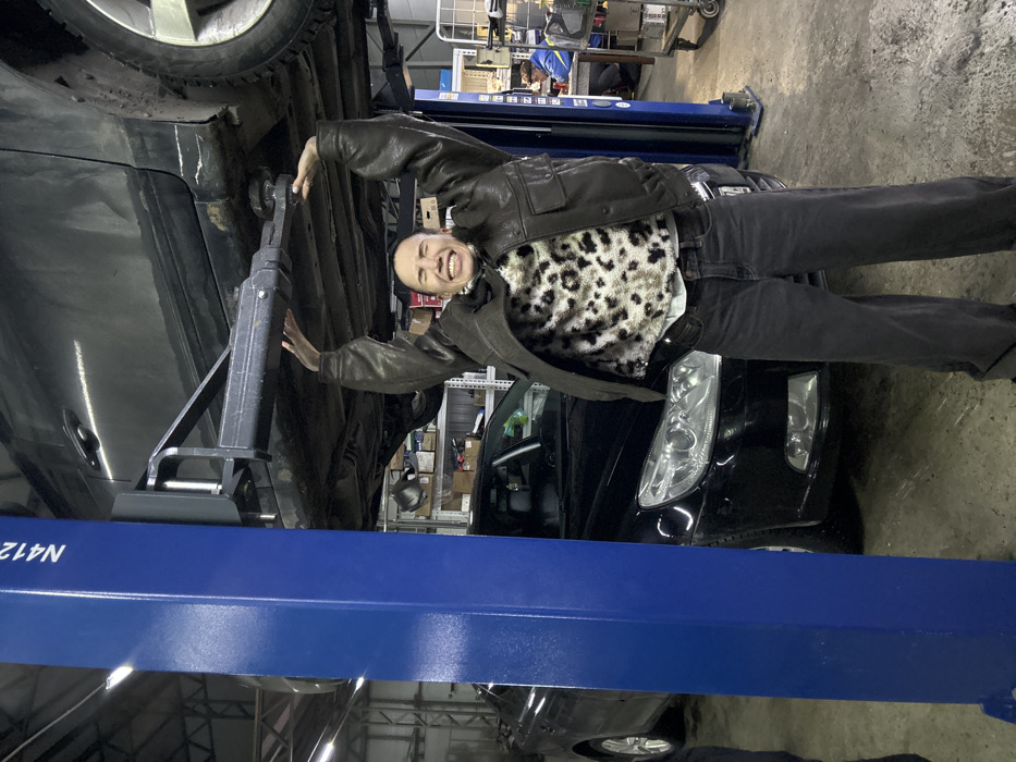 The image captures a vibrant scene inside what appears to be an auto repair shop or garage. The primary subject is a person, positioned prominently in the center of the frame, smiling broadly with an open mouth, showcasing their teeth. They have short hair and are dressed in a dark brown or black leather-like jacket worn over a light-colored, possibly cream or white, shirt featuring a distinctive leopard print pattern with dark spots. They are also wearing dark pants, likely jeans, and dark shoes. The person is standing upright, facing the camera, with their arms slightly bent and hands extended towards the underside of a vehicle. Their left hand appears to be resting near a support mechanism under the car.
Two cars are visible in the scene. One dark-colored vehicle, possibly an SUV or crossover, is elevated high on a hydraulic car lift, with its underside and one of its wheels (in the upper left) clearly visible. Below and to the right of the lifted car, another dark-colored car, whose front end including headlights and grille is visible, is parked on the ground.
A large, robust blue hydraulic car lift forms a significant part of the setting, with its pillars supporting the elevated vehicle. On one of the blue support structures, the text "N412N" is visible. A black metal car jack or support stand is positioned directly under the lifted car, close to the person's left hand, providing additional stability.
The background reveals typical garage elements, including shelving units stocked with various items and tools, suggesting a functional workspace. The flooring appears to be concrete, and the overall lighting indicates an indoor, well-illuminated environment. The person's wide smile and energetic pose convey a sense of excitement or playfulness, making the image feel dynamic and engaging.