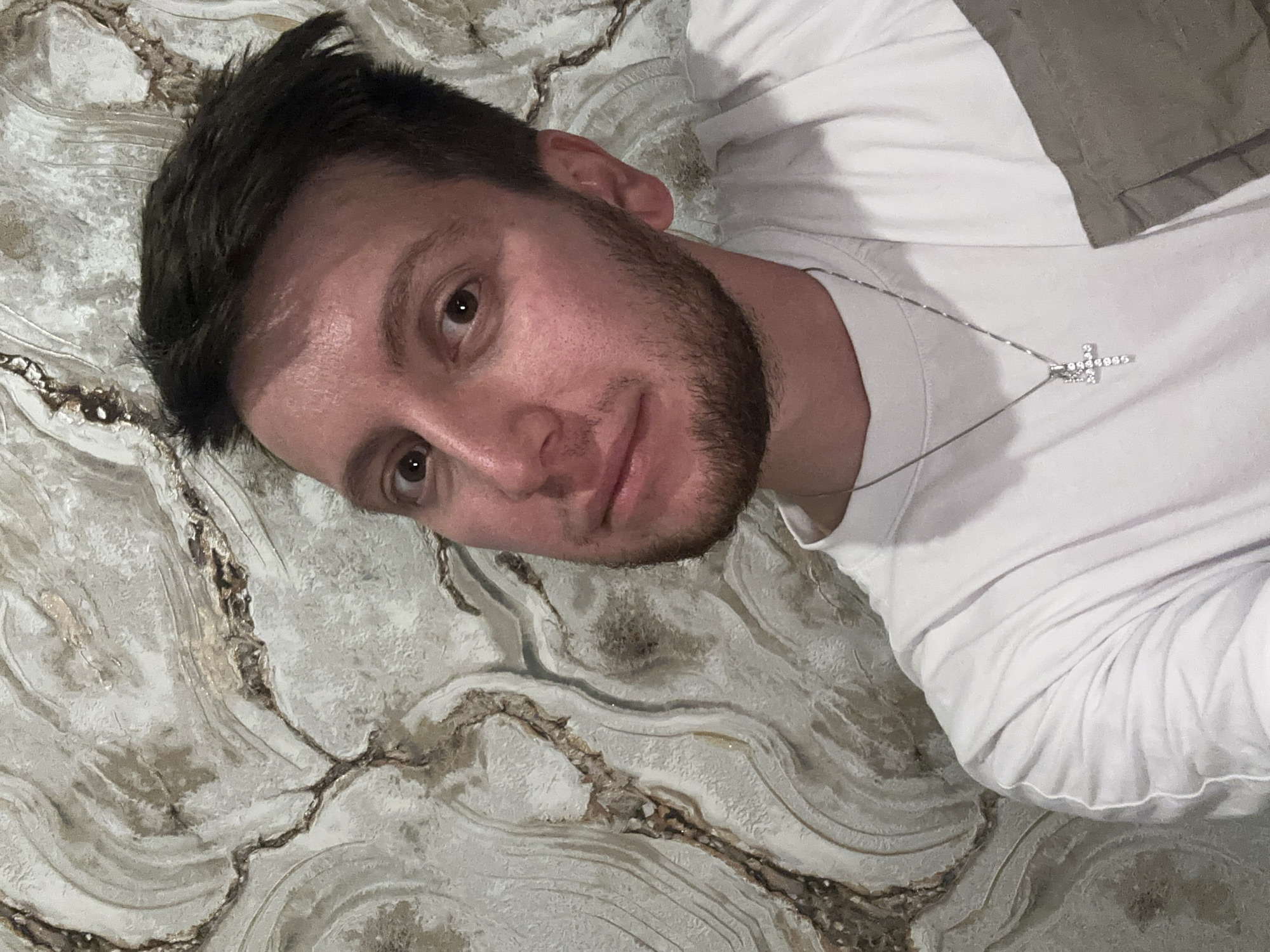 A medium shot shows a young Caucasian man with short, dark brown hair and a light beard, looking directly at the camera. He is wearing a white t-shirt with a silver cross necklace. The man is positioned in front of a wall with a marble-like pattern in shades of grey and beige. The background is detailed with organic, swirling lines. The image is taken from the chest up, with the man's face slightly angled. The lighting is somewhat dim, casting subtle shadows on his face. There is no identifiable information regarding the specific location within the city of Mirny, Russia.