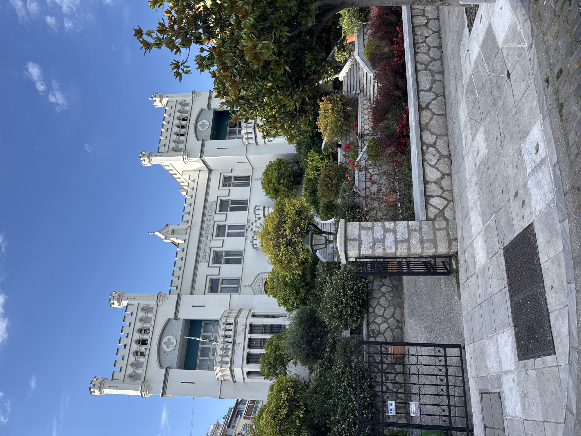 The image shows a grand, white, castle-like building with a crenellated facade and turrets. Above the entrance, the Greek word "ΔΗΜΑΡΧΕΙΟΝ" (Dimarchion, meaning Town Hall) is inscribed. The building is surrounded by lush greenery, including trees, shrubs, and flowering plants, and a stone wall. A black wrought-iron gate stands open in the foreground, leading to a pathway and stairs. The sky is blue with scattered clouds. The scene appears to be a public building in a well-maintained urban area.