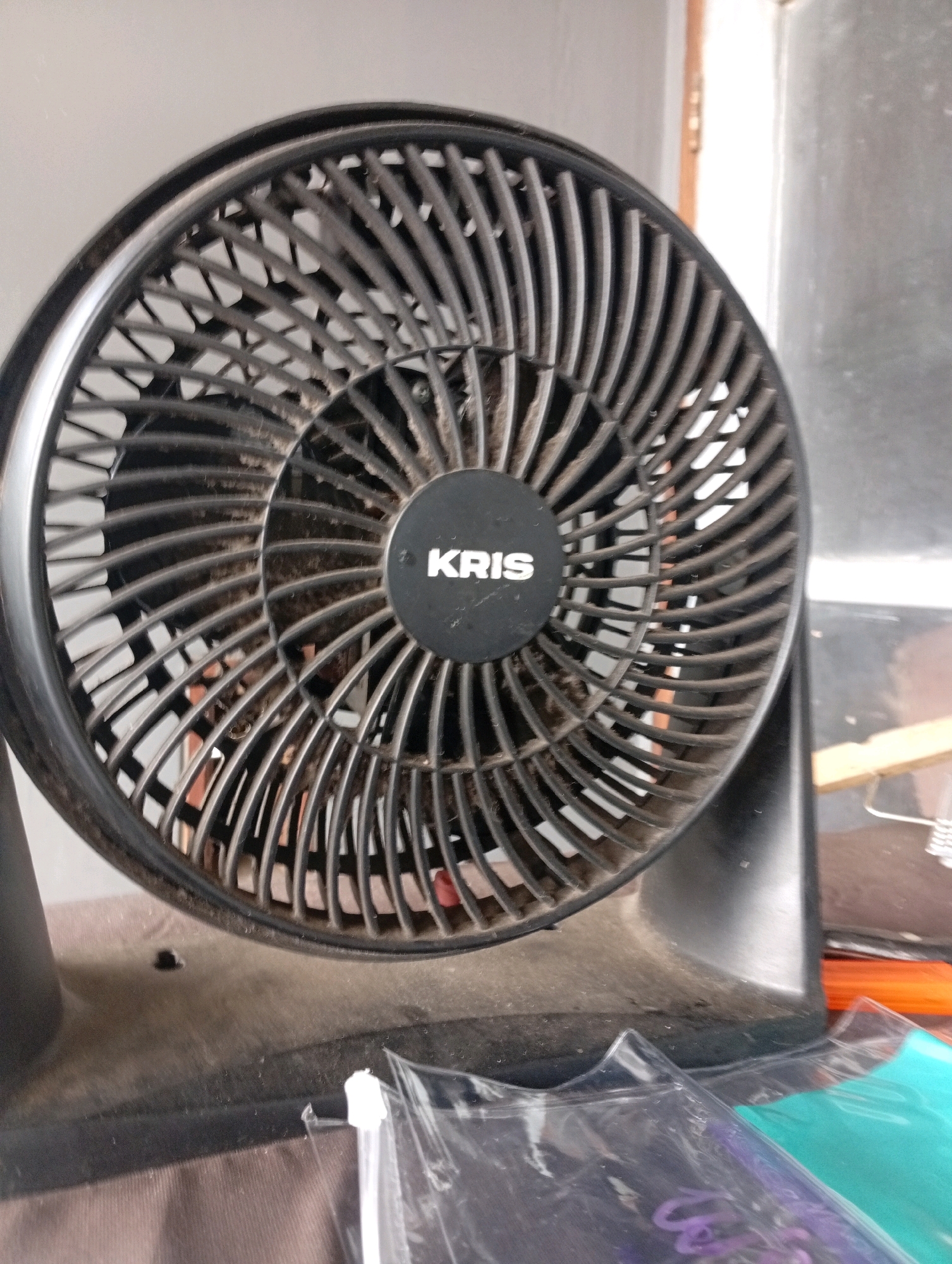 The image shows a close-up of a dusty black electric fan with the brand name "KRIS" visible on a circular emblem in the center of the fan grille. The fan is positioned on a surface that appears to be a table or desk, with other objects, including some clear plastic packaging with purple and teal colored items inside, visible in the foreground. The background is blurred but suggests an indoor environment with a wall and a doorway. The lighting is natural, coming from the right side of the image. The overall impression is of a domestic or office setting, likely in Tasikmalaya, Indonesia, given the context provided.