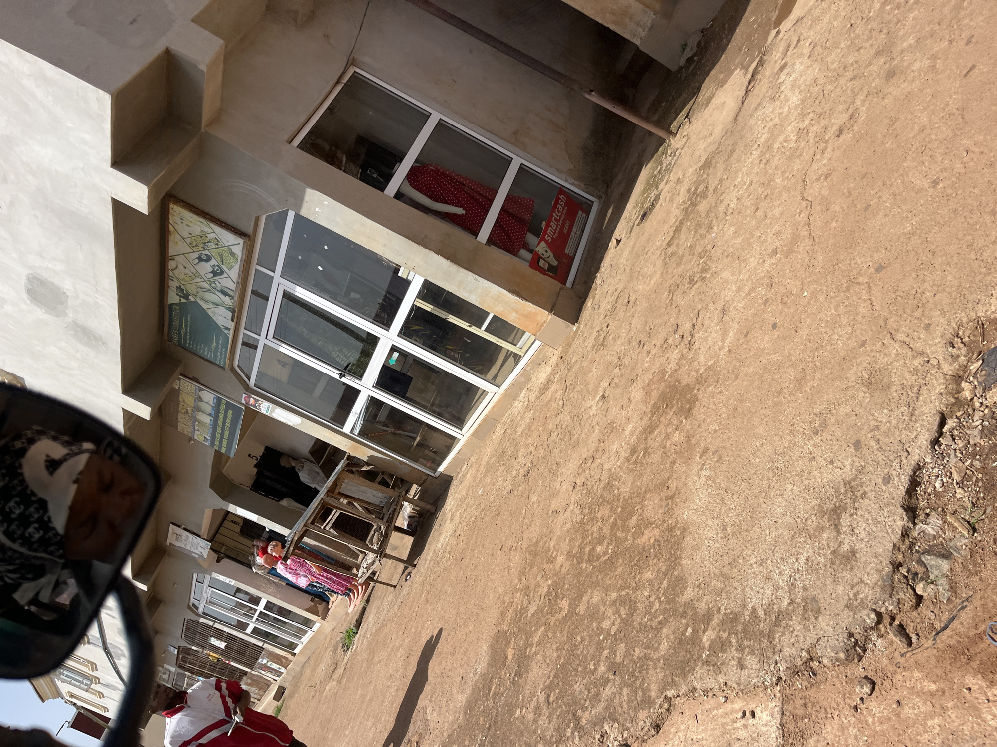 The image depicts a street scene in Gura Top, Nigeria, with shops lining a dusty road. A man wearing a red and white jacket is standing near the left side of the frame, facing away from the camera. In front of the shops, mannequins dressed in colorful clothing are displayed. One mannequin is wearing a red and white polka dot dress. A poster advertising "smartcash" is visible in the window of one shop. The buildings appear to be made of concrete, with large glass windows and metal security bars. The ground is covered in dirt and gravel. The sky is visible in the upper portion of the image, appearing bright and clear. A reflection in a mirror shows a person's face wearing a patterned headscarf.
