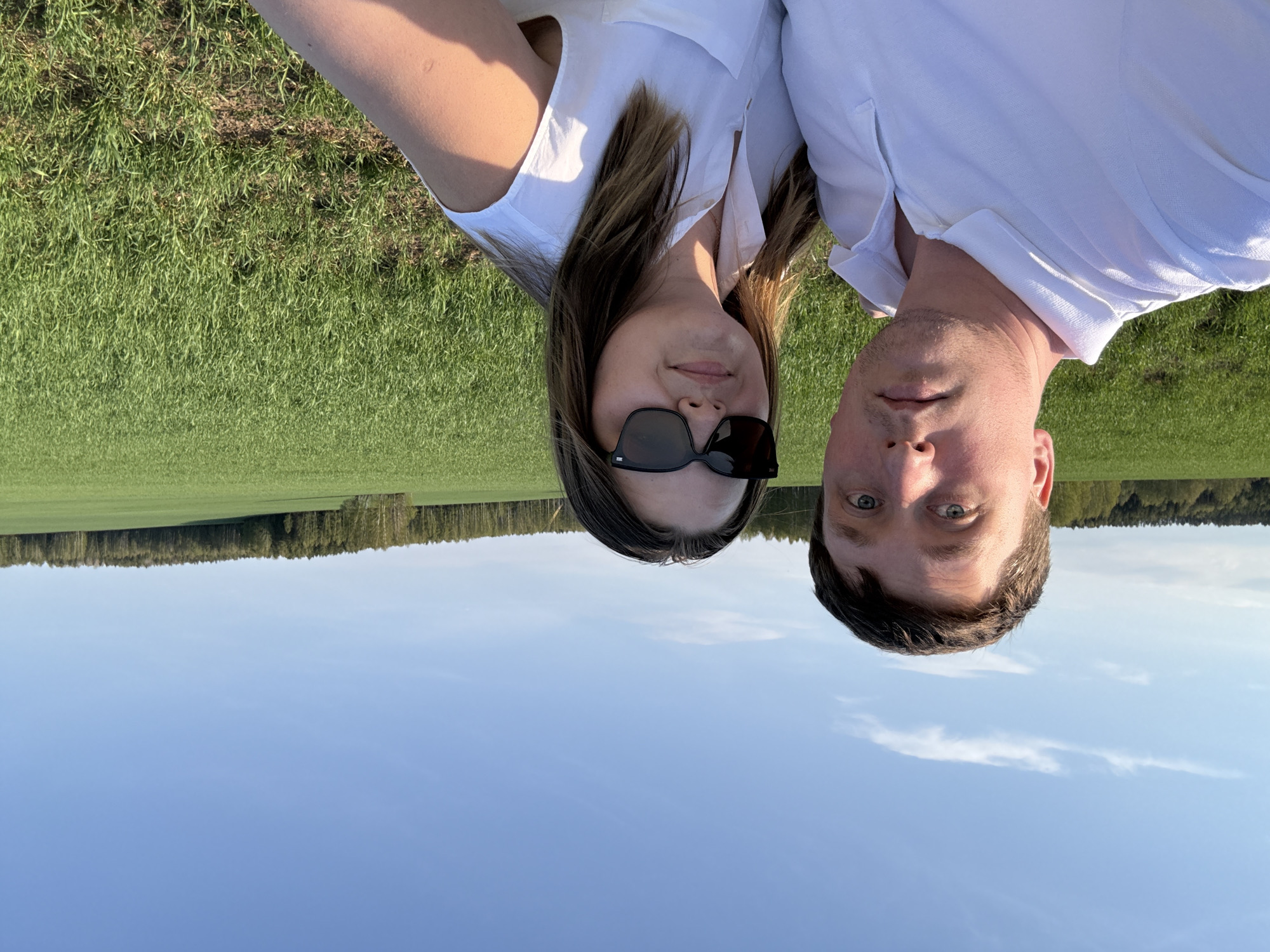 A selfie features a man and woman, both wearing white shirts, against a background of green fields, a treeline, and a blue sky. The image orientation is inverted, making the sky appear below and the fields above. The woman wears sunglasses. The location is presumed to be near Шумиха, Russia, based on contextual information.