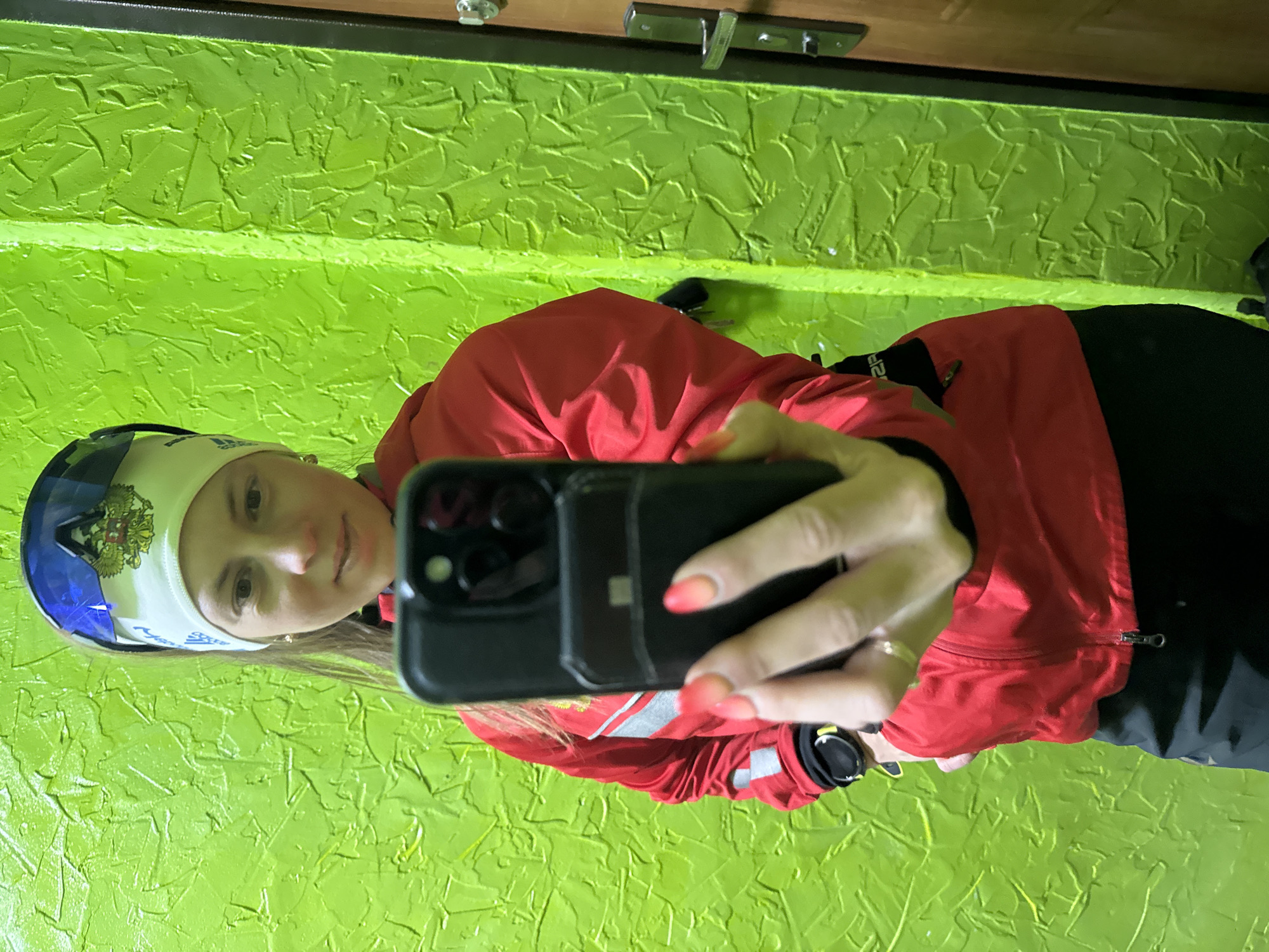 A woman takes a selfie with a smartphone. She is wearing a red jacket, black pants, and a white headband with a blue design and Russian coat of arms on a blue background. She is positioned in front of a green textured wall, likely indoors. The location is in Chaikovsky, Russia, is indicated by the coat of arms. A doorknob is visible in the background.