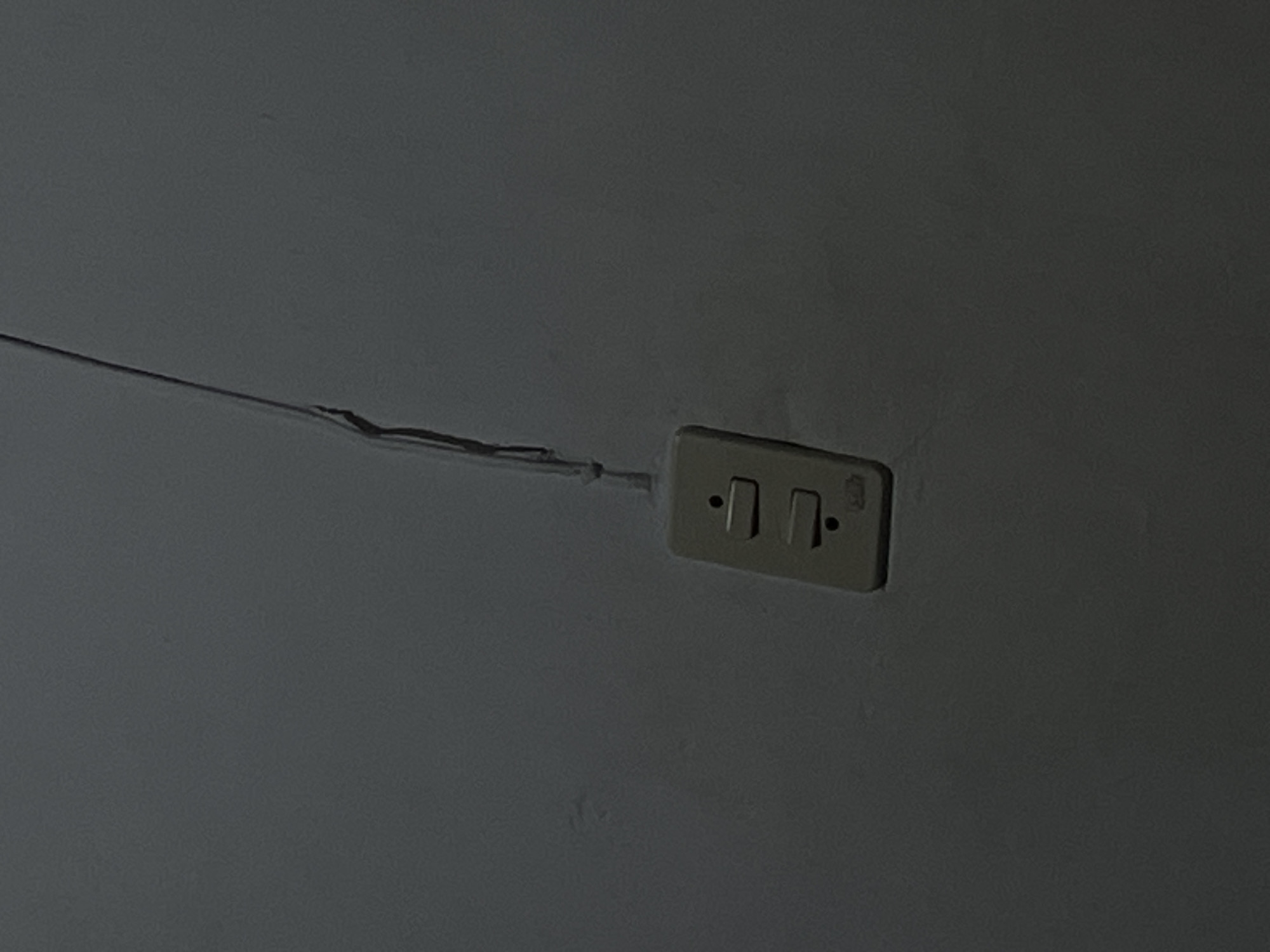 This media file shows a close-up, eye-level shot of a white electrical outlet with two toggle switches. The outlet is mounted on a plain, gray wall. A thin, black electrical wire runs diagonally across the wall from the left side of the frame, disappearing towards the outlet. The lighting is dim, casting a shadow and creating a muted color palette. The overall impression is one of simplicity and a focus on the functional object. There is no visible text in the image.