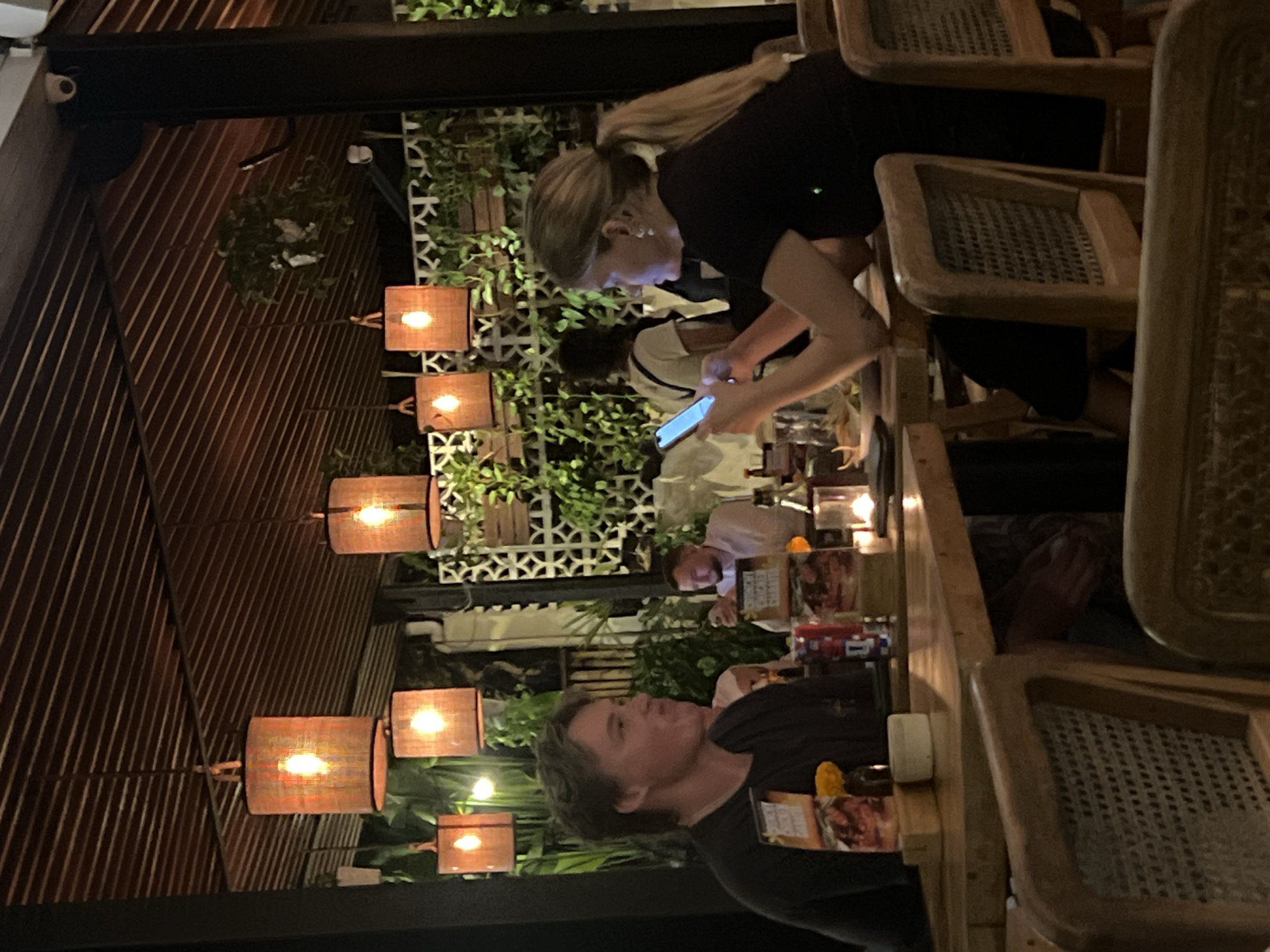 The image depicts an outdoor dining scene at a restaurant in North Kuta, Indonesia. A woman with blonde hair pulled back in a ponytail is standing at the table, looking at and interacting with a smartphone. She is wearing a black top and has a visible tattoo on her arm. Another person at the table is also using a smartphone, while a man in a black t-shirt looks towards the camera. There are multiple people seated at the table. The table is set with food, condiments, and candles. Wicker chairs surround the table. The environment is decorated with hanging lights with wicker shades, and a wall covered in plants and a geometric pattern provides a backdrop. The lighting suggests it is nighttime. The architecture features wood slats. A security camera is visible in the upper left corner.