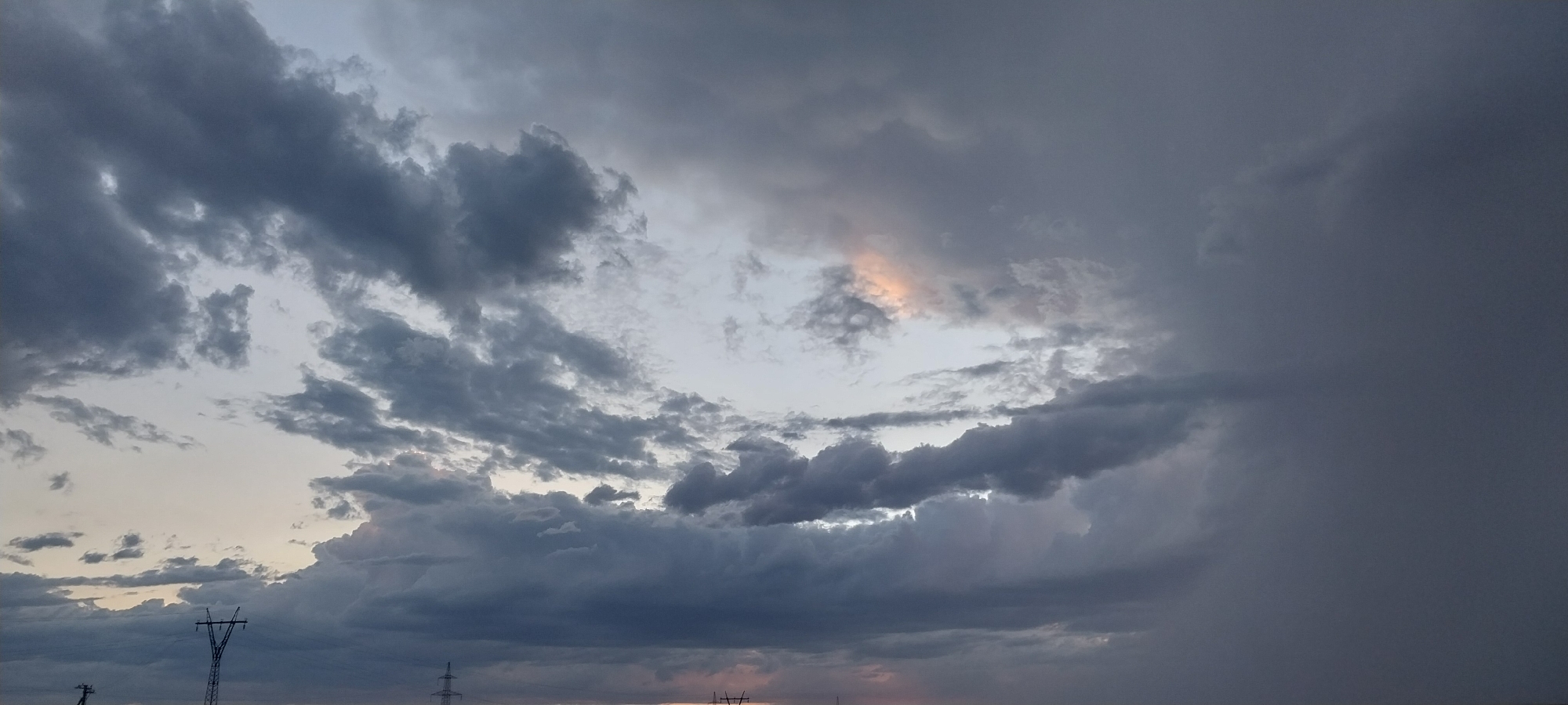 An image captures a dramatic sky filled with dark, brooding clouds, suggesting an impending storm. The clouds vary in texture and shade, with some appearing wispy and others dense and heavy. A small patch of light pinkish-orange hue breaks through the gloom in the upper center of the frame, offering a contrast to the otherwise somber tones. The lower portion of the image shows the silhouettes of two power line towers against the twilight sky. The left tower is clearly visible, a tall lattice structure with wires extending from it. The right tower is partially obscured and less distinct. The overall scene conveys a sense of atmospheric tension and the power of nature. The location is listed as Unknown, Unknown.