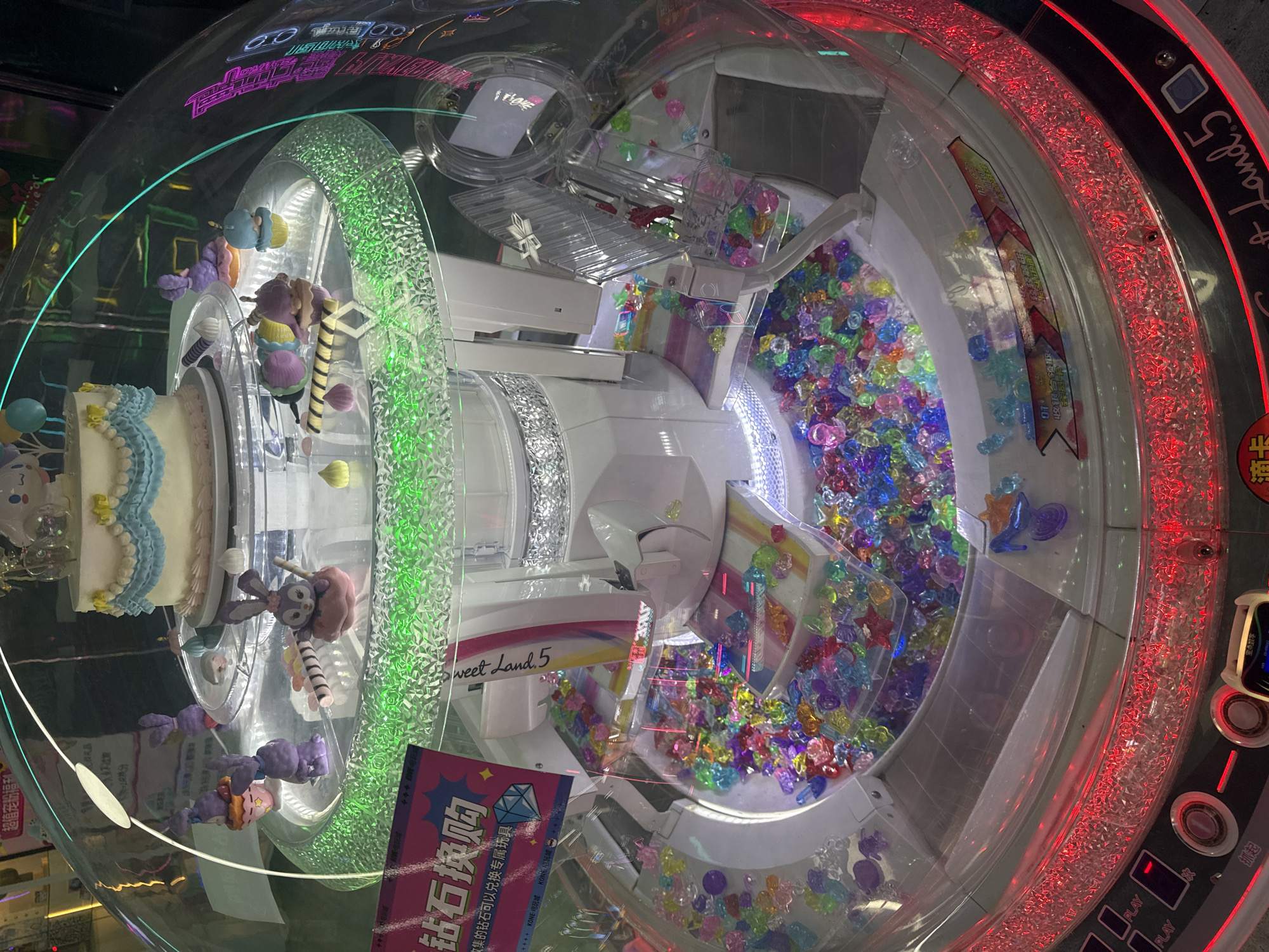 The image shows a close-up view of a colorful arcade prize machine, likely a claw or prize dispensing game, filled with numerous plastic gems and small decorative items. At the top of the machine, a tiered cake-like structure is visible, adorned with blue and yellow icing and topped with a character resembling a cloud. Several small purple bunny figurines are placed around the cake on a transparent platform illuminated with green lights. The lower section of the machine contains a large quantity of brightly colored, faceted plastic gems in various shapes, resembling cut jewels. A sign in Chinese characters, which translates to "Diamond Exchange," is partially visible on the left side of the machine, suggesting a reward system. The machine itself is enclosed in a clear plastic dome, and the base features red LED lighting and control buttons. The scene is likely located in a commercial setting such as a shopping mall or entertainment center in Yiwu, China, a city known for its vast wholesale markets and vibrant commercial activity. There are no people visible in the image, and the focus is entirely on the prize machine and its contents. The overall impression is one of a playful and visually stimulating arcade game.
