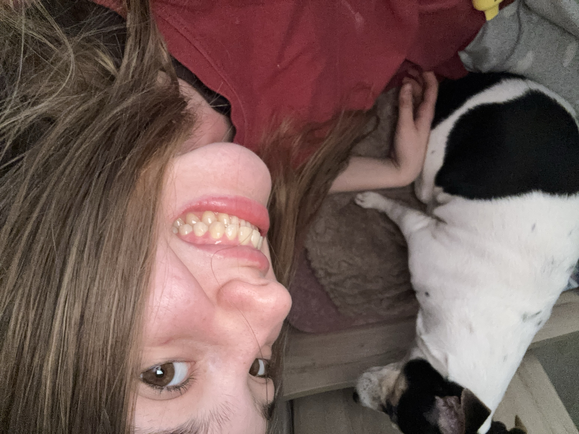 The image is a close-up, selfie-style photograph featuring a person and a dog. The person, likely a young female, is positioned in the foreground, with their face tilted towards the camera. They have brown hair and are wearing a red top. Their mouth is open, revealing teeth with braces. To the left of the person, a small black and white dog is lying down, partially visible. The background appears to be an indoor setting, with what looks like wooden furniture and a textured surface. The lighting is dim, casting a warm tone over the scene. No specific location in Vsevolozhsk, Russia is discernible from the image content.
