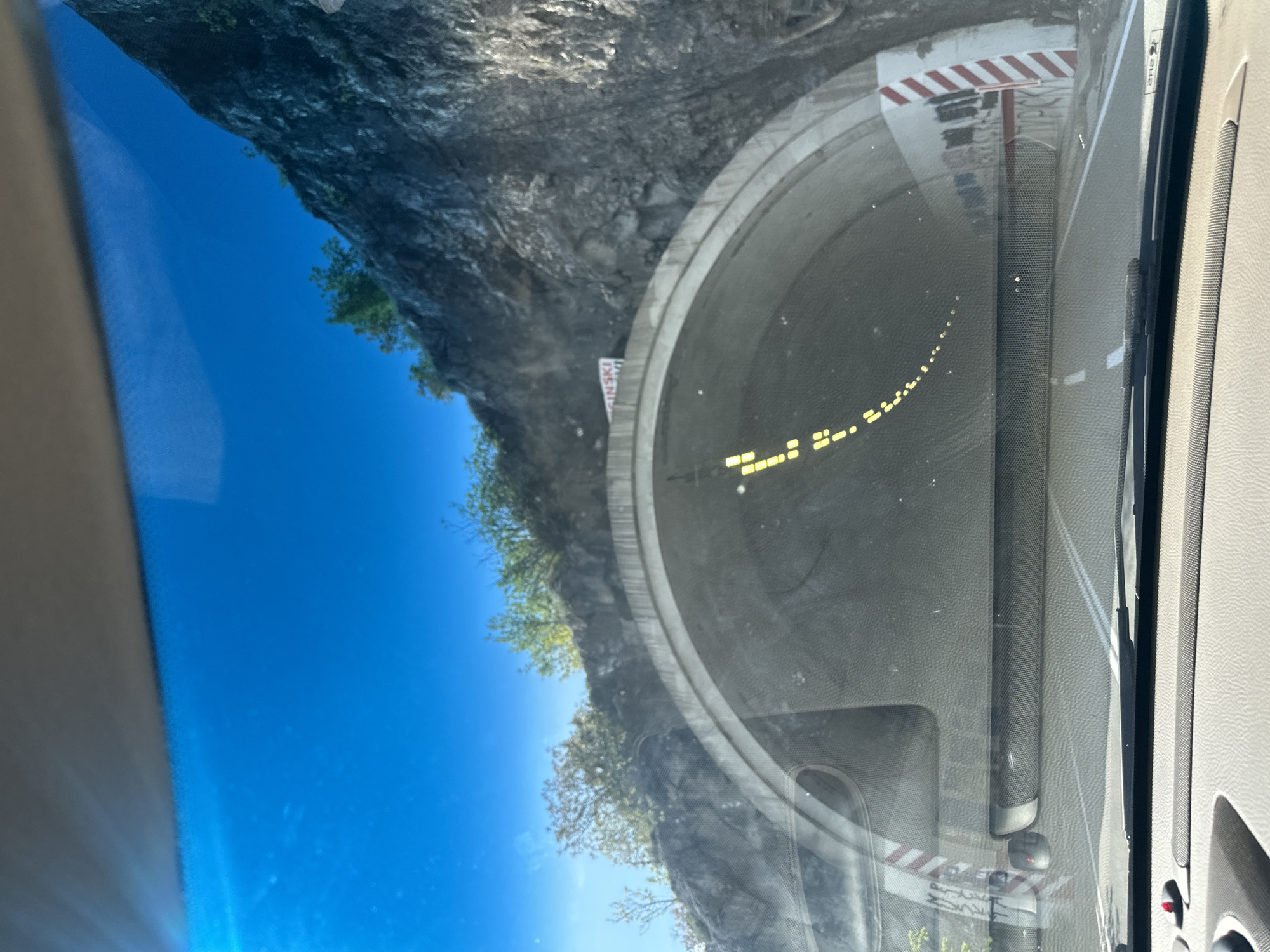 The image, taken from inside a vehicle, captures the moment of approaching a tunnel through a rugged, mountainous landscape in Strugari, Montenegro. The view is through the car's windshield, with reflections visible across its surface.
In the center, a large, grey concrete tunnel entrance dominates the scene. Inside the tunnel, a series of bright yellow, reflective markers are visible along the left wall, guiding the path. To the left of the tunnel entrance, a steep, rocky cliff face rises, partially covered with sparse green trees and vegetation. Above, a clear, vibrant blue sky indicates a sunny day.
Reflected prominently in the windshield on the right side is a red and white striped barrier or gate, possibly indicating a road closure or warning at the tunnel entrance. Also visible through the windshield is a white sign near the tunnel entrance, with the partial text "GINSKI" and a letter "M" or "MT" below it. The car's interior, likely a dashboard or pillar made of light grey material, is visible along the right and bottom edges of the frame. The overall impression is of a car traveling on a road winding through a scenic, rocky terrain under clear weather conditions.