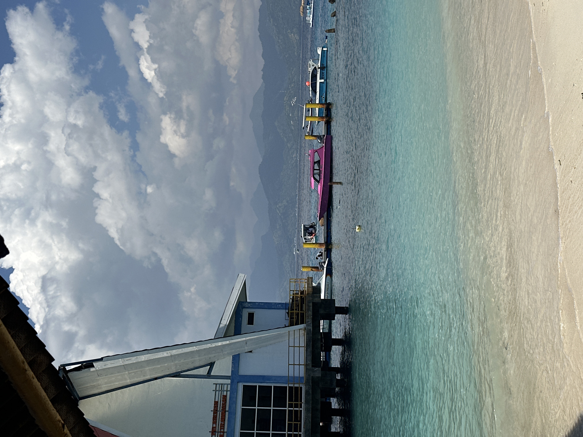 The image captures a coastal scene in an area of Unknown, Indonesia, featuring a dock extending into clear turquoise water. The dock supports several boats, including a notable pink one, indicating transport or recreational marine activity. The shoreline shows a sandy beach adjacent to the water. Adjacent to the dock is a building with a white and blue facade. The background reveals mountainous terrain under a sky with mixed cloud cover.
