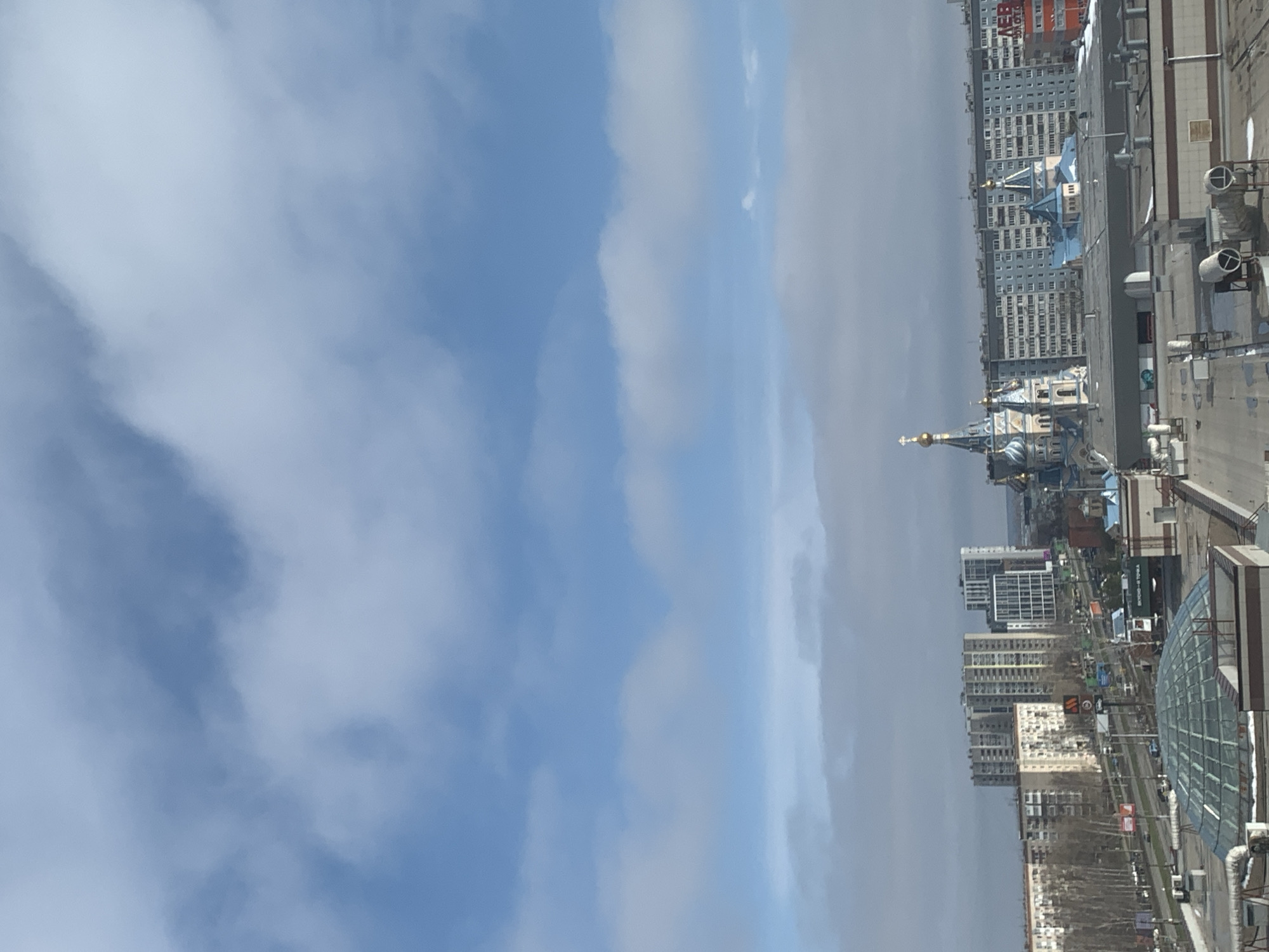 The image depicts a cityscape in Izhevsk, Russia, likely viewed from an elevated position. The sky is filled with clouds. Visible are buildings of varying heights, a church with blue domes and a golden spire, rooftop structures, and possibly streets with minimal activity.