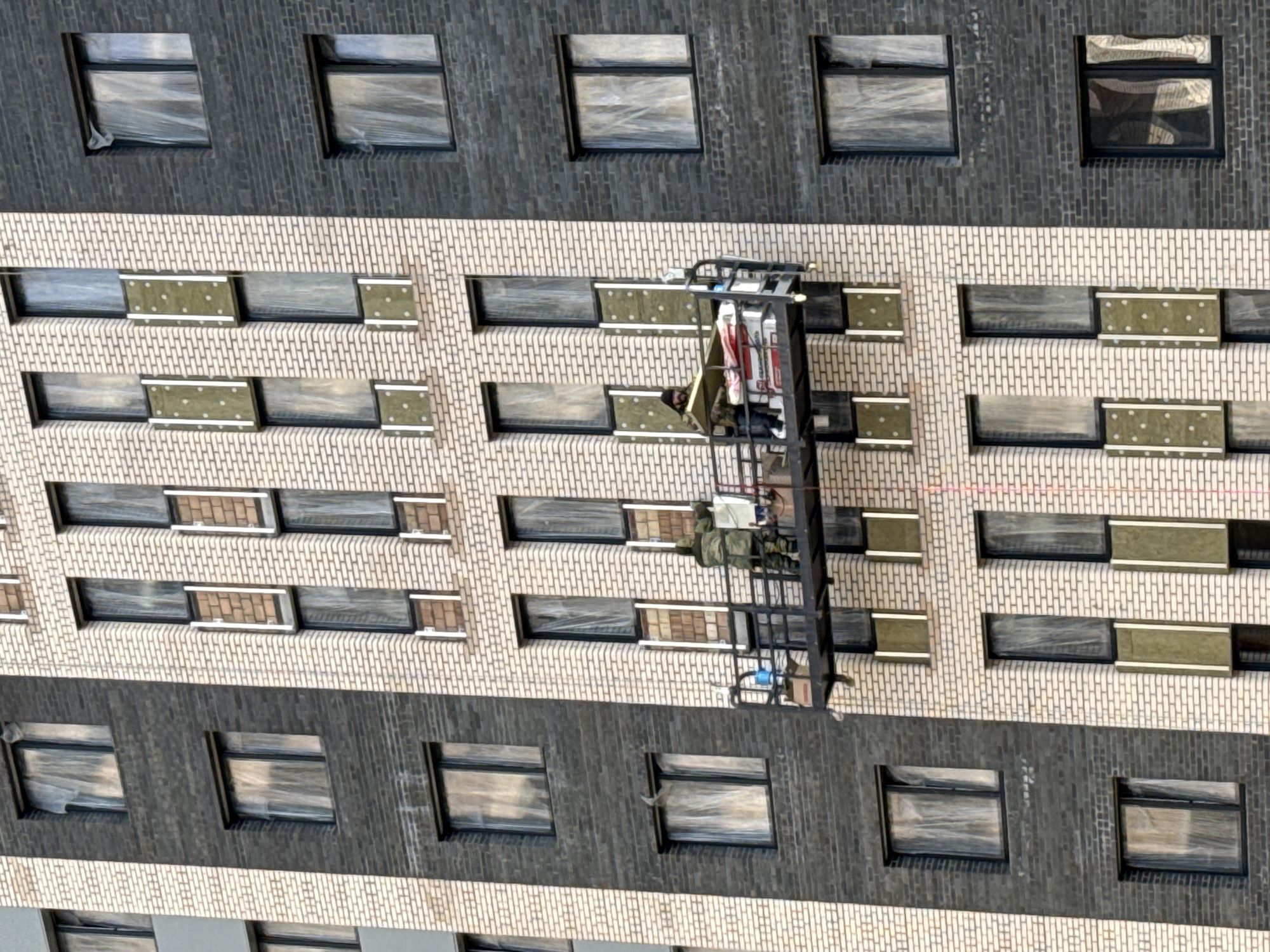 The image shows the exterior of a multi-story building, likely in the city of Rusanovo, Russia. The building has a repeating pattern of windows across multiple floors. Some windows appear to be boarded or covered with a material, while others are open. A mobile lift platform is positioned against the building, with one or two workers visible. The lift contains various items, including clothing and tools. A red caution line runs across the facade of the building. The building is constructed of brick, with alternating sections of light and dark-colored bricks. The overall scene suggests construction, renovation, or repair work is in progress.