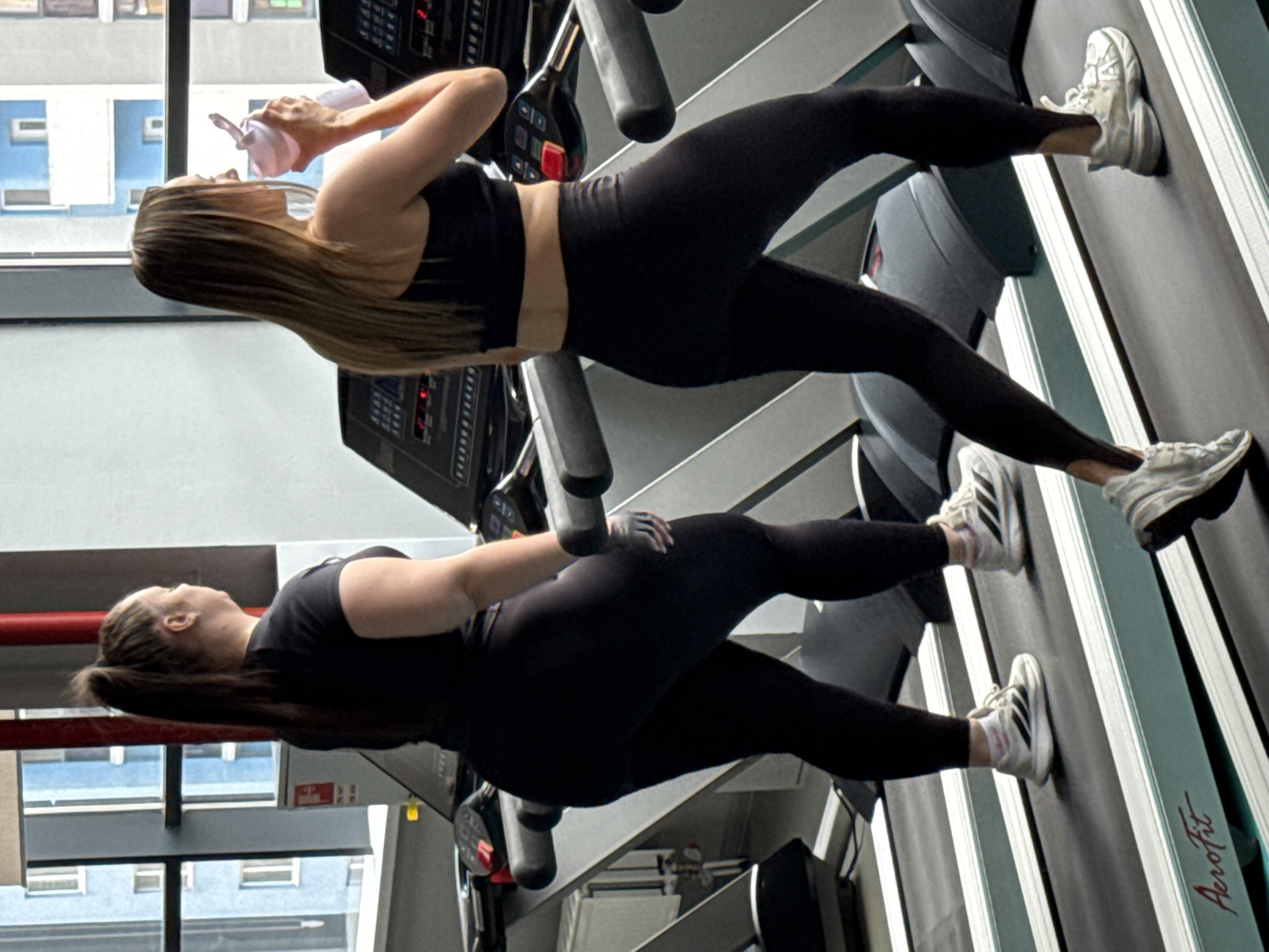 This image captures two women engaged in exercise on treadmills within a gym setting in Yekaterinburg, Russia. The photo is rotated 90 degrees clockwise, showing the subjects vertically within the frame.
The woman in the upper part of the image has long, light brown hair and is dressed in a black sports bra and black leggings. She is actively drinking from a pink water bottle, suggesting a pause during her workout. Her feet, clad in white athletic shoes, are visible on the treadmill belt.
The second woman, positioned lower in the frame, has dark brown hair tied back in a ponytail. She is wearing a black t-shirt and black leggings, and a black workout glove can be seen on her right hand. Her white sneakers, featuring black stripes, are also on the treadmill belt.
Both women are utilizing treadmills equipped with control panels and handrails. Through a large window in the background, the exterior of urban buildings can be seen, providing a glimpse of the city outside. The brand name "Aerofit" is distinctly visible on the side of one of the treadmills. The scene depicts a moment of fitness activity in a gym.
