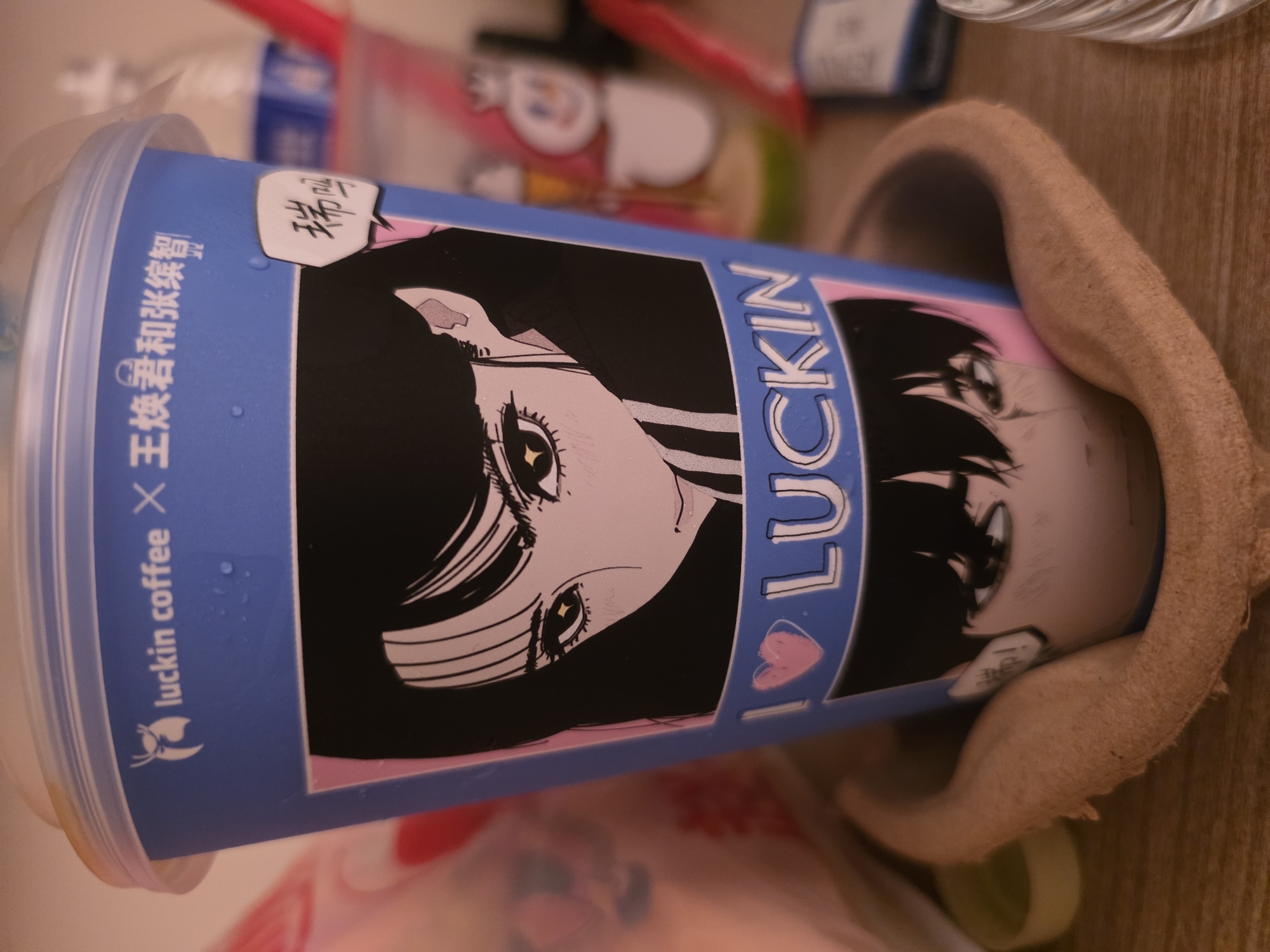 The image shows a blue Luckin Coffee cup with anime-style artwork. The top of the cup has the text "luckin coffee" and some Chinese characters. Below this, there is an illustration of a woman's face with dark hair and large eyes, followed by the text "I LUCKIN" with a pink heart. The bottom part of the cup shows the lower half of a man's face. The cup is placed in a tan, textured cup holder on a wooden surface. In the background, out of focus, are other cups and objects, suggesting an indoor setting. There is no discernible information about the location being in Luolong District, China.