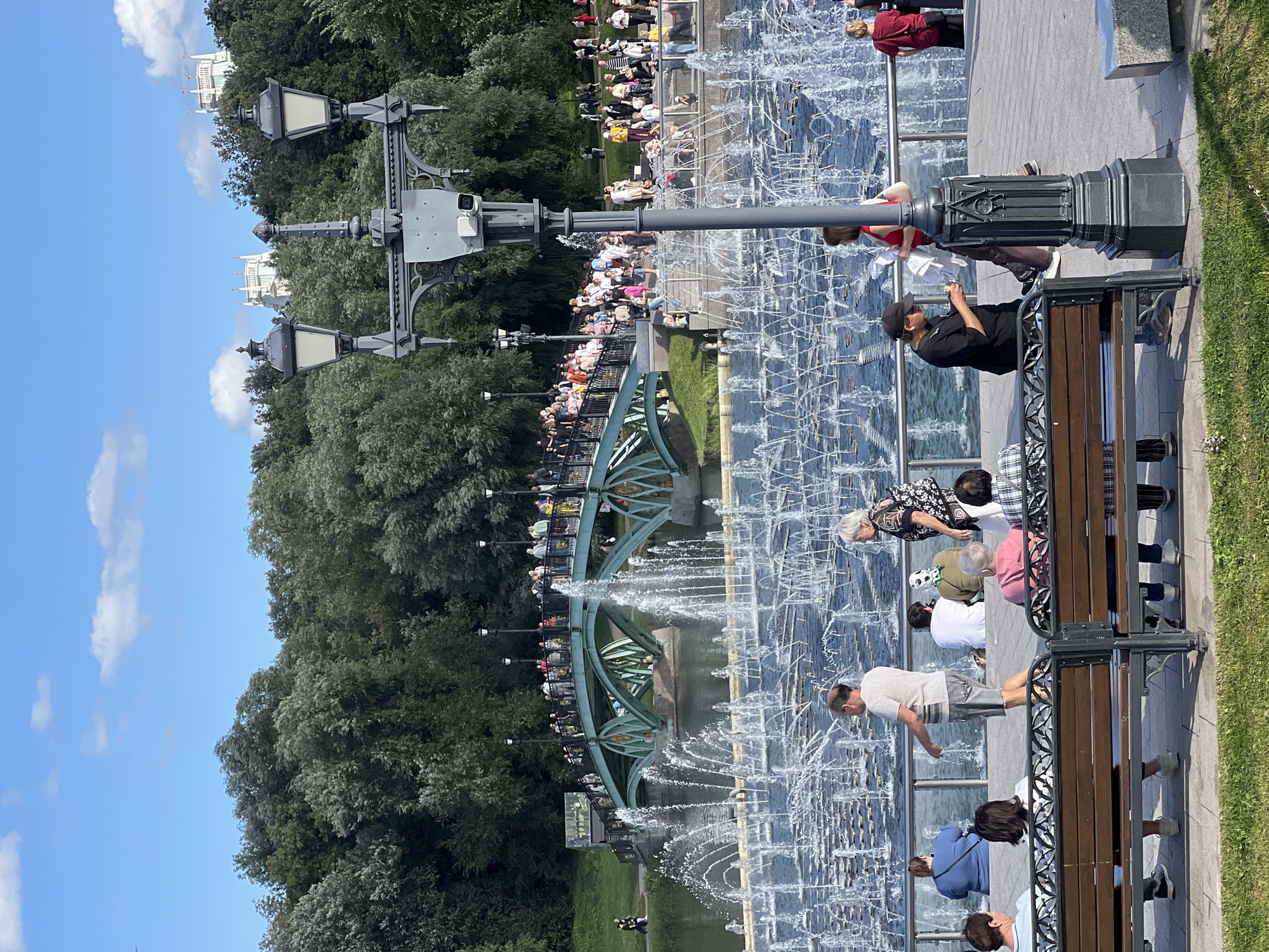 The image depicts a sunny day scene at Tsaritsyno Park in Moscow, Russia. In the foreground, several people are seated on benches, facing away from the camera, observing a large, multi-tiered fountain with numerous water jets. A lamppost with two lamps stands prominently in the middle ground, with a security camera attached to its base.
In the background, a distinctive green arched bridge spans a body of water. A crowd of people is gathered on and around the bridge, suggesting it is a popular spot. Behind the bridge, a dense forest of green trees forms a lush backdrop against a clear blue sky with scattered white clouds. In the far distance, on a hilltop, light-colored architectural structures, possibly part of the Tsaritsyno Palace complex, are visible amidst the trees. The overall atmosphere is one of a public park on a pleasant day, with people enjoying the water features and the scenery.