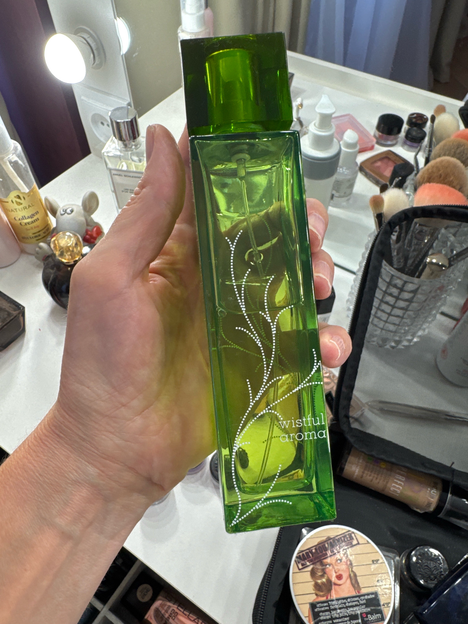 A hand holds a tall, rectangular, translucent green glass perfume bottle with a matching green cap. The bottle has a white vine-like design etched on its surface and the text "wistful aroma" is visible near the bottom. The hand is positioned in the foreground, obscuring part of the bottle.
In the background, on a white surface, are various cosmetic items. To the left, a small clear bottle with a gold label reads "NATURAL Collagen Cream." Next to it is a small, grey, mouse-shaped figurine with red accents. Further back, a silver-capped perfume bottle is visible.
To the right of the perfume bottle, a black makeup bag is open, revealing an assortment of brushes and other makeup products. A clear container with brushes and what appears to be glitter is nestled inside.
The environment suggests a vanity or makeup station, with a mirrored surface and lighting fixtures visible. There is no discernible connection to the city of Sinyavino, Russia. The image is a close-up shot focusing on the perfume bottle and the hand holding it.