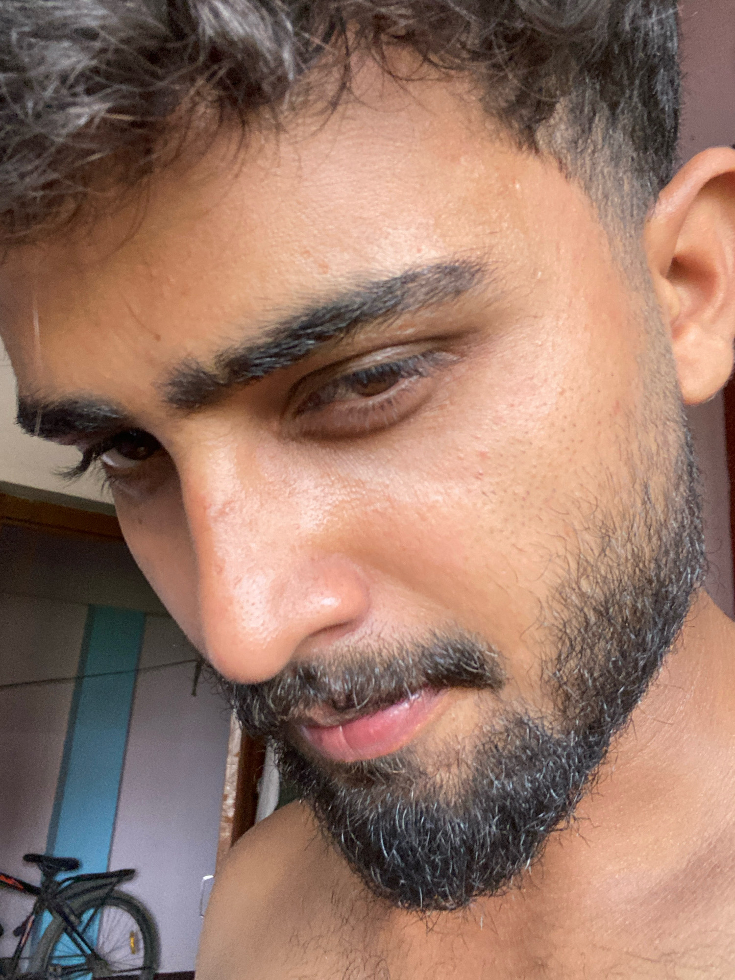 The close-up, medium-shot, eye-level photograph captures a person's face, focusing on the right side of their face including their eye, nose, mouth, and beard. The person has dark hair styled with a fade cut and a thick, salt-and-pepper beard. Their right eye is open and looking down and to the left, with visible eyelashes. The skin appears slightly oily and shows some texture. The background is blurred, but suggests an indoor or semi-outdoor setting with a wall that has vertical blue and grey stripes and a portion of a bicycle on the left. The lighting suggests natural light coming from the right. The image was taken in Bahadurpur, India.