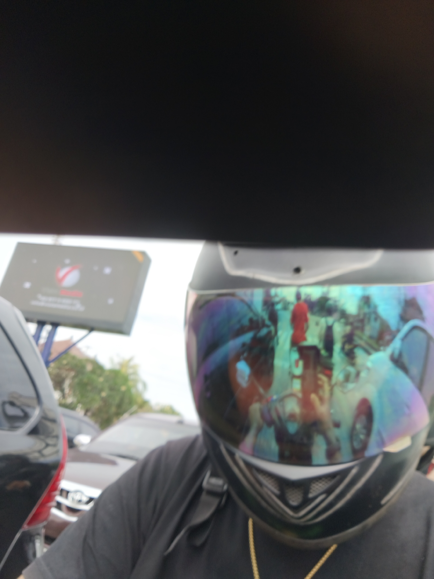 A selfie taken from the perspective of someone wearing a black t-shirt and a black motorcycle helmet with a iridescent visor. The visor reflects a busy street scene with several vehicles, including cars and motorcycles. One car is a dark color, possibly black or dark brown, and another car appears to be a lighter color. A person in a bright red shirt is visible, and other figures are present in the reflection. In the background, a large, rectangular billboard with a logo featuring a red and white swoosh is mounted on a blue structure. Trees and foliage are also visible behind the billboard, suggesting an outdoor environment. The overall scene appears to be a street in Denpasar, Indonesia, given the provided location context. The image is somewhat blurry, particularly the reflections and background elements.