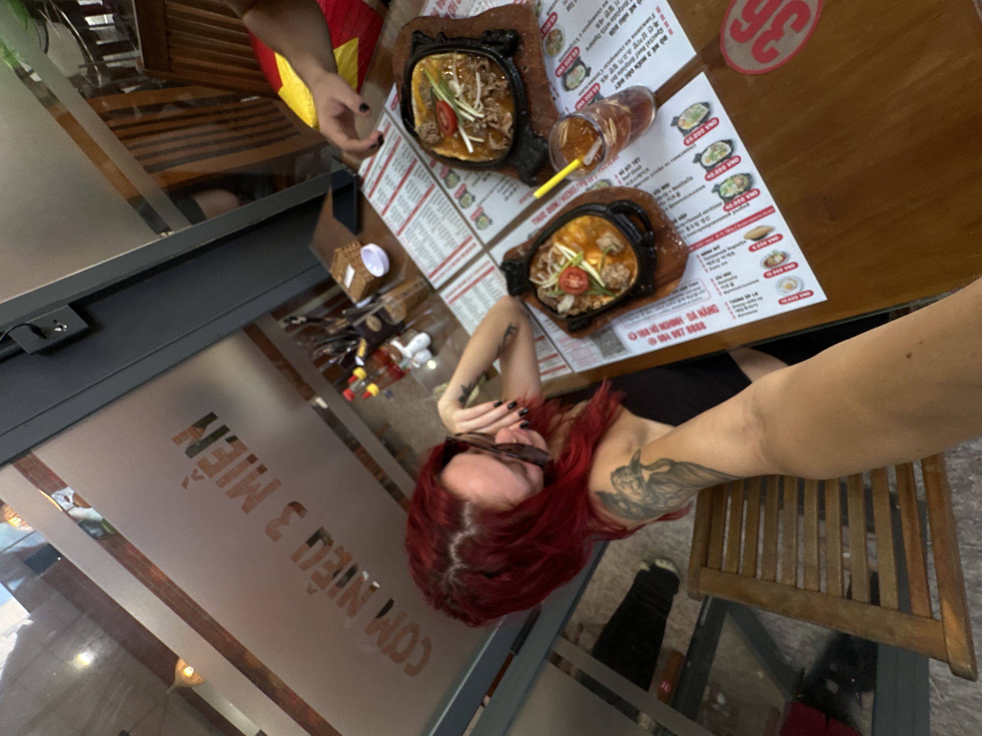 A young woman with bright red hair and tattoos on her arm is seated at a table in a restaurant in Da Nang, Vietnam. She is wearing sunglasses and a black top. In front of her are two sizzling platters of food, one with meat and vegetables, and another with what appears to be meatballs and broth. A glass of iced tea with a yellow straw sits on the table, next to a menu. The restaurant appears to be casual, with wooden chairs and tables, and frosted glass windows. The menu is in Vietnamese and appears to have prices listed in Vietnamese Dong. A portion of the restaurant's signage, "CƠM NIÊU 3 MIỀN", is visible on the frosted glass window. The woman is taking a selfie, with her arm and part of her face visible in the foreground. The overall scene suggests a meal at a local eatery, likely a popular spot given the menu and the food presentation.
