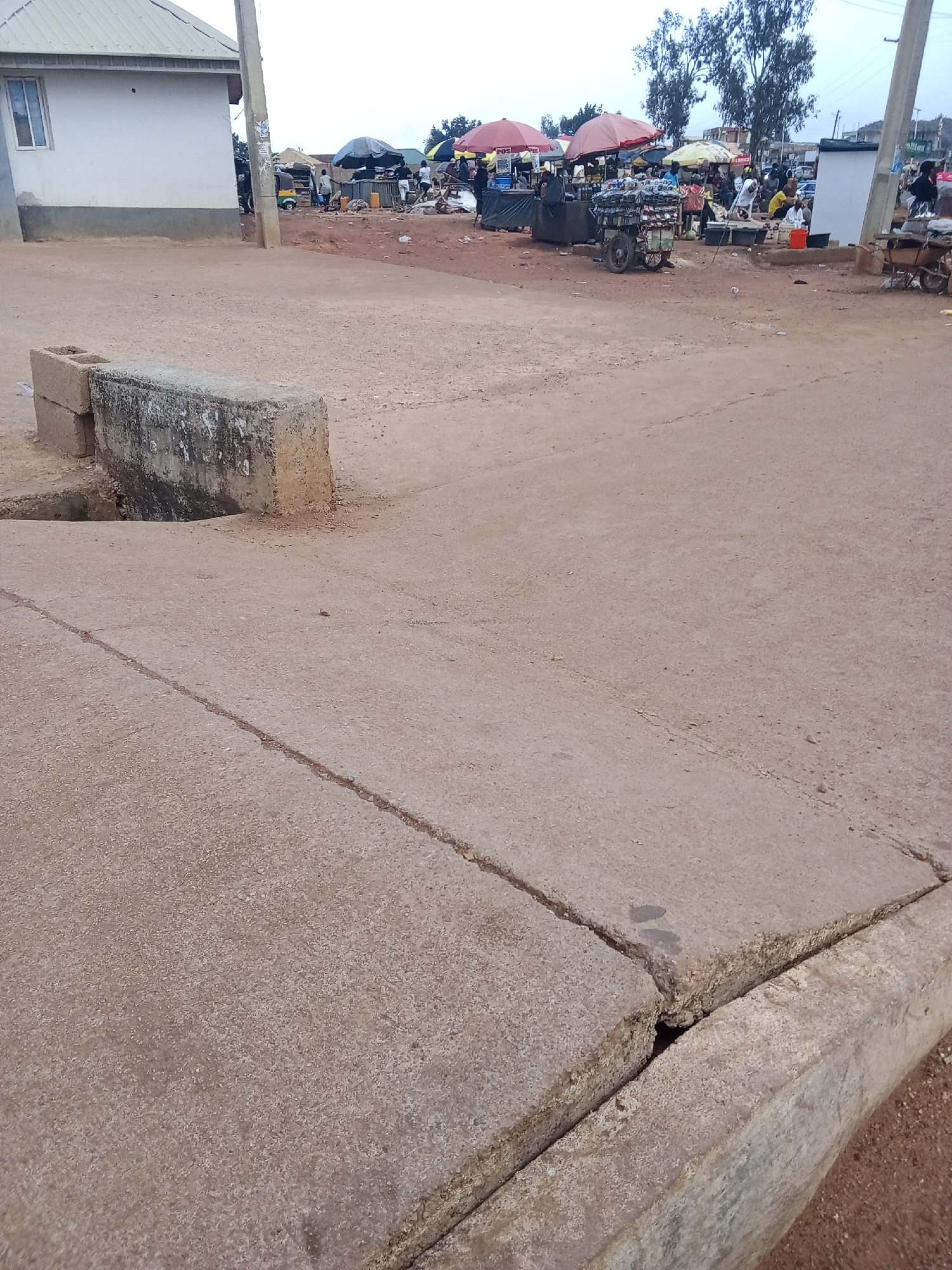 The image depicts a street scene in Gura Top, Nigeria, likely a marketplace. In the foreground, a cracked concrete surface is visible, with a concrete block barrier to the left. In the background, stalls with red and yellow umbrellas are set up, with people milling about. Some of the stalls appear to be selling goods, including what looks like packaged items on a cart. Several individuals are standing or sitting around the stalls, and a motorized tricycle, possibly a "keke napep," is parked among them. The environment appears dusty and unpaved, with trees visible in the distance and a building with a white facade to the left. The sky is overcast.