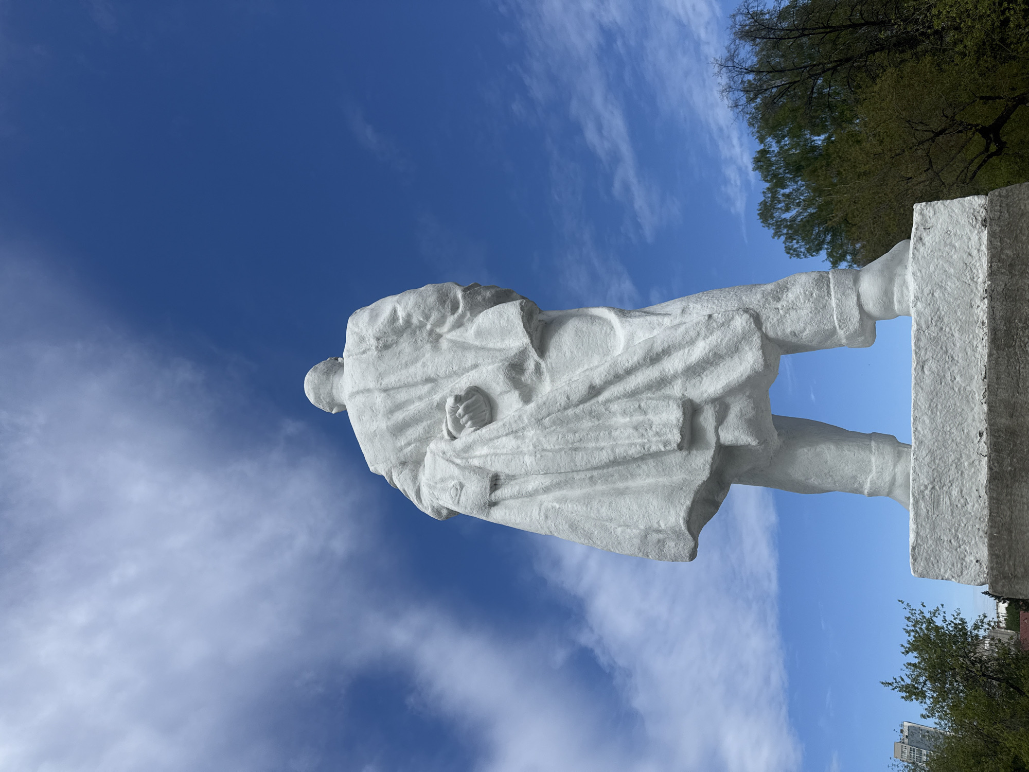 The image shows a white stone statue of Vladimir Lenin, the founder of the Russian Communist Party. The statue depicts him in a long coat, holding a fist. The statue is located in Yekaterinburg, Russia, likely in a public square or park. The sky is bright blue with scattered white clouds. Trees are visible in the background. The photo is taken from a low angle, looking up at the statue.