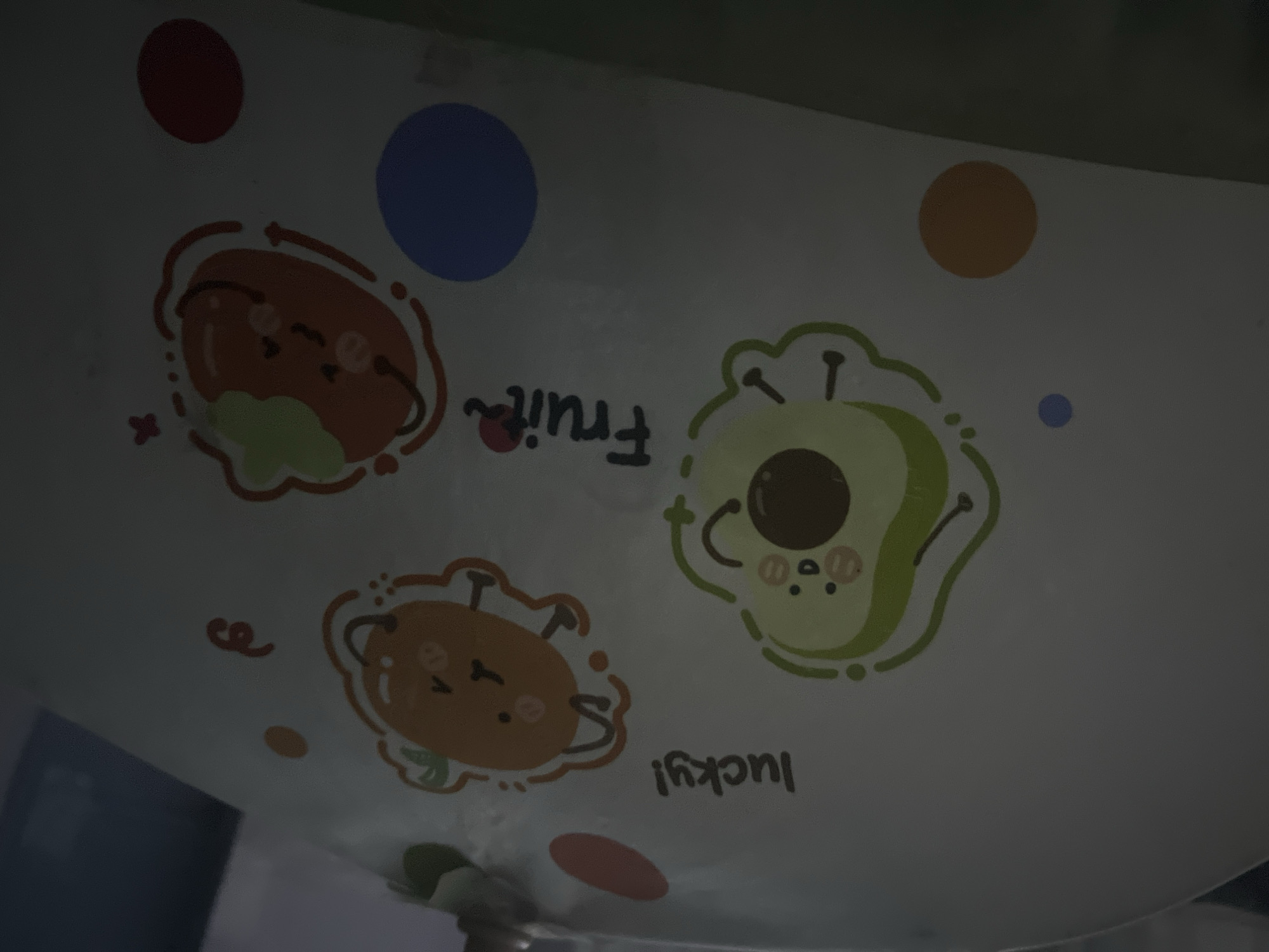 A dimly lit interior scene shows a close-up of a white surface with cartoonish drawings of fruits and text. In the upper left, the word "lucky!" is written in black cursive. To the right of the text, an orange fruit with a smiling face and arms is depicted. Below the text and to the left, a green avocado with a smiling face and arms is drawn. In the center, the word "Fruit" is written in black cursive with a red dot above the "i". To the right of the word "Fruit", another orange fruit with a green top, smiling face, and arms is illustrated. Scattered around the drawings are various colored dots, including orange, blue, and red. The lighting is low, casting shadows and making details in some areas difficult to discern. This image is not located in Chong-Sary-Oy, Kyrgyzstan.