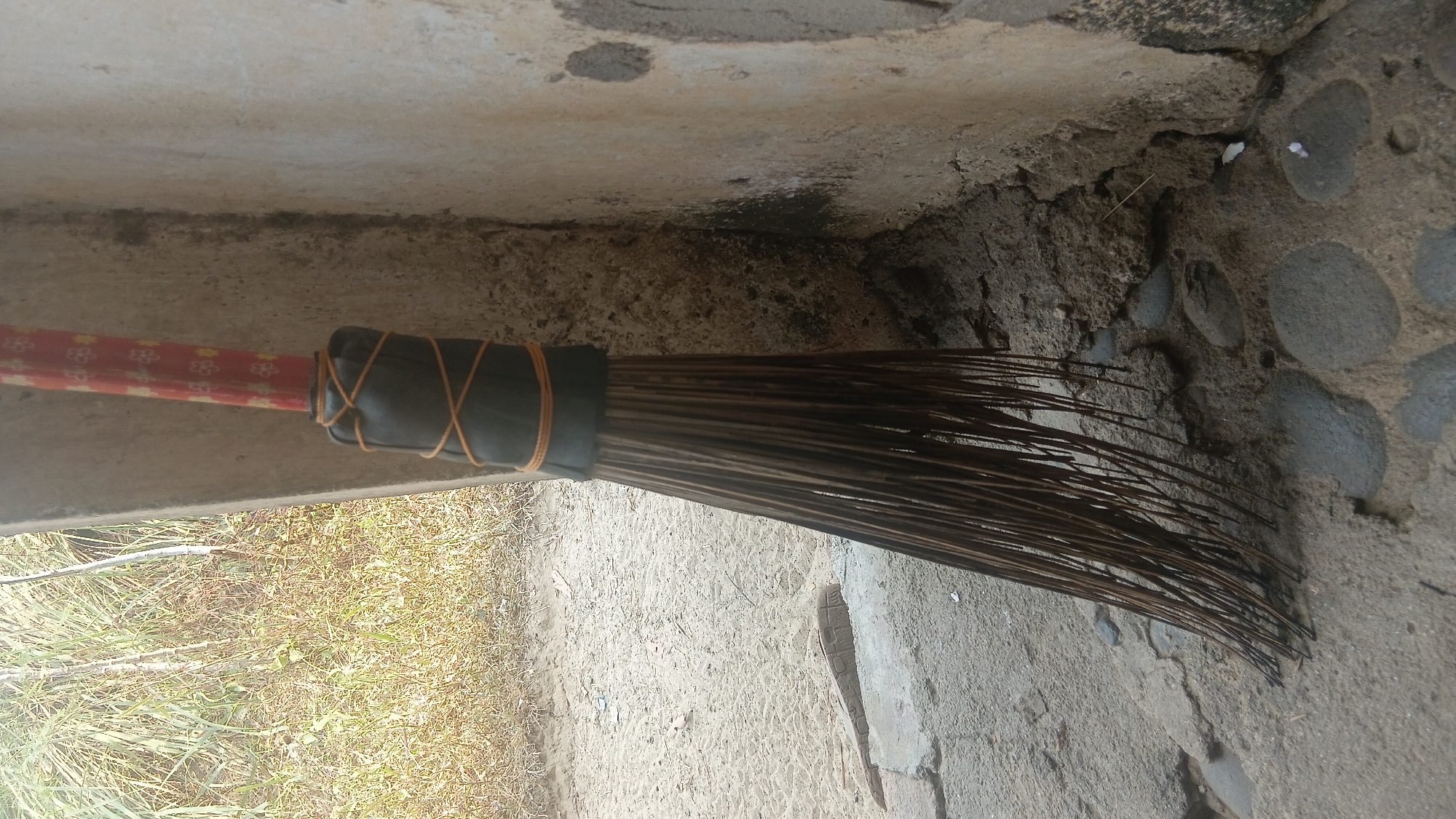 An outdoor, ground-level, medium shot captures a broom leaning against a textured, light-colored concrete wall. The broom has a red handle decorated with a repeating pattern of yellow flowers. The lower part of the handle is wrapped with black material and secured with crisscrossed orange cord. The broom's bristles are dark, thin, and appear to be made of natural fibers, fanned out at the bottom. The ground in front of the wall is a mix of rough concrete and dark stones. To the left of the broom, a sandy area with dry grass is visible. The lighting suggests it is daytime, with some shadows cast on the wall. There are no people or animals present in the image. The overall scene depicts a utilitarian object in an outdoor, possibly slightly neglected, urban or semi-urban environment. This scene could be found in various locations within Prabumulih, Indonesia, such as near a residential dwelling, a shop, or a public pathway.