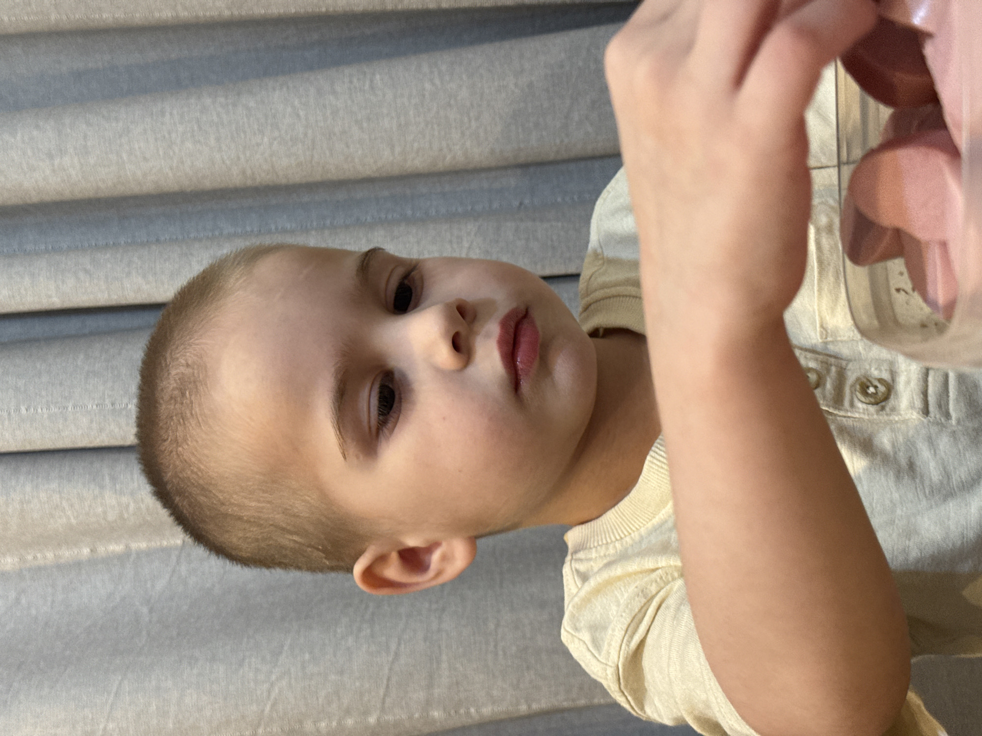 A single juvenile individual is depicted indoors in Chaikovsky, Russia, within a private residential setting. The specific location within the city is not discernible from the image. The juvenile, appearing male, has light skin, dark eyes, and very short, light brown hair. He wears a light-colored, short-sleeved Henley shirt with button details. His gaze is directed downwards and to the right. His left arm and hand are visible reaching into a clear plastic container in the lower right of the frame, which holds multiple pink, heart-shaped objects, possibly candies or toys. The background consists of a light gray or muted blue-gray curtain with vertical folds.