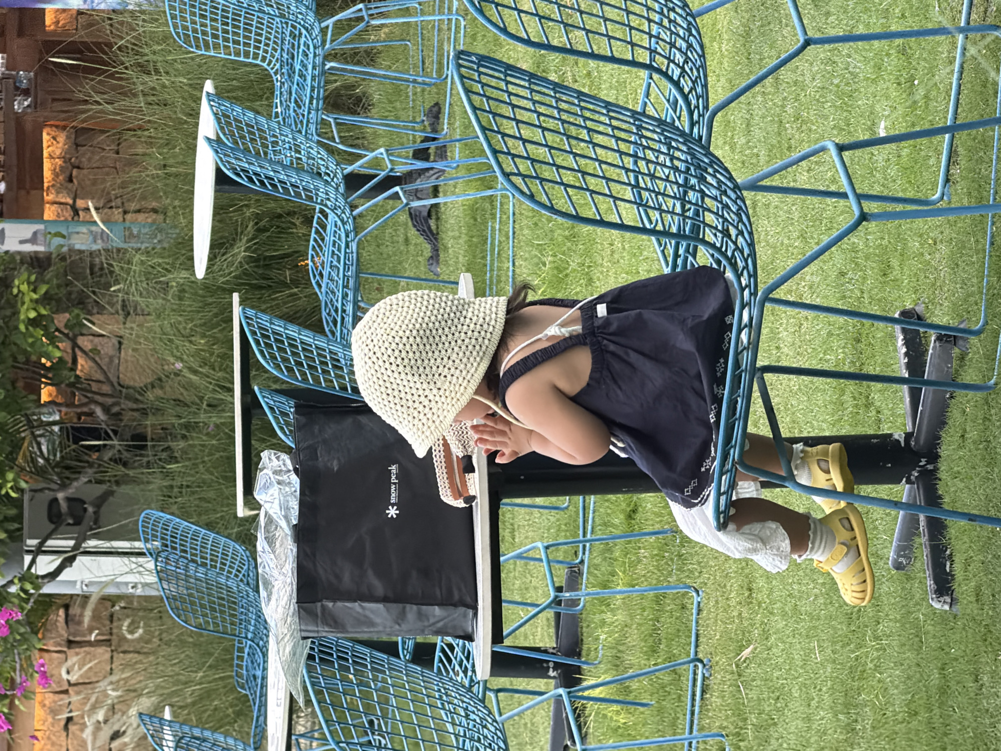 The image depicts a child sitting alone at an outdoor table in what appears to be a cafe or restaurant setting. The child, facing away from the camera, wears a dark dress, a light-colored hat, white socks, and yellow sandals. They are seated on a blue metal chair and their hands are clasped in front of their face. On the table is a black bag with "snow peak" written on it, along with some crumpled plastic and a patterned object. The surrounding environment consists of more blue metal chairs and tables, a patch of green grass, and in the background, a stone wall and foliage. Based on the available information, this scene is likely located at the Sailing Club Nha Trang, a popular beachfront venue in Nha Trang, Vietnam, known for its distinctive blue chairs and outdoor seating area.