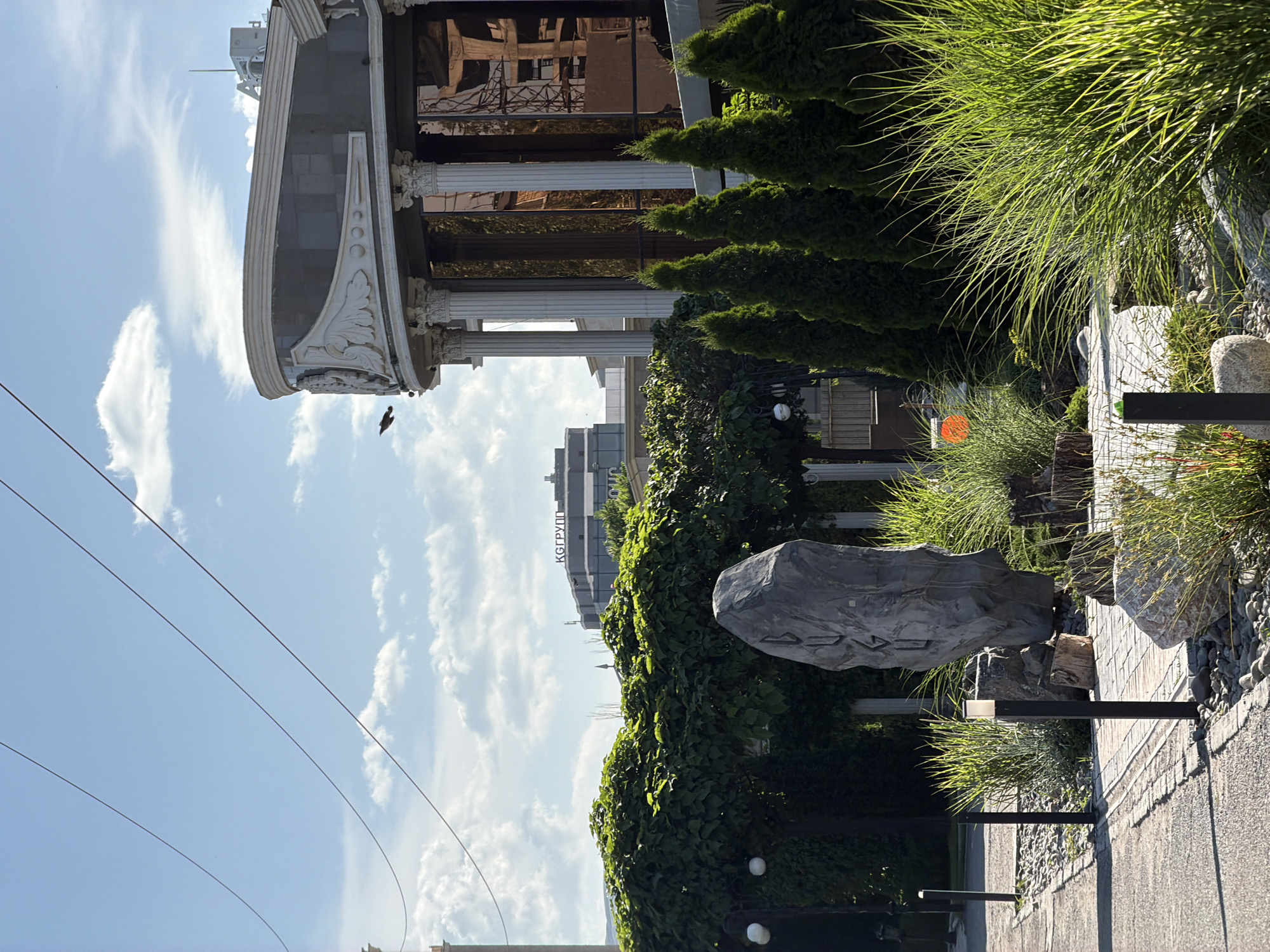The image captures an outdoor scene in Bishkek City, Kyrgyzstan. The foreground features a paved walkway alongside a landscaped area with rocks, plants, and decorative lighting. A large rock with some kind of inscription is prominently displayed. Lush greenery, including shrubs and tall grasses, is abundant. In the background, there is a building with Greek-style columns and architectural details, reflecting other structures in its windows. Further back, a taller building with the word "КГГРУПП" is visible. Overhead power lines stretch across the blue sky with scattered clouds. A bird is in flight.