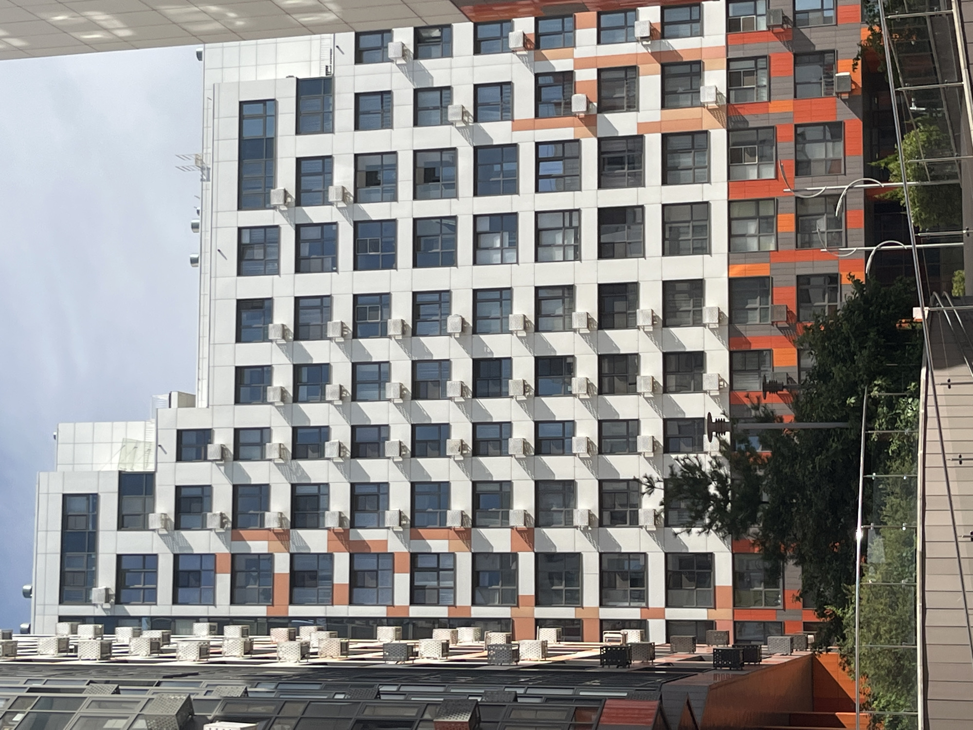 This is a medium-angle, eye-level shot of a modern residential building in Moscow, Russia, likely part of a new development in the Izmaylovo district, given its architectural style and recent construction. The building itself is tall and multi-story, with a white facade interspersed with repeating patterns of dark grey and bright orange square panels. Numerous windows, each with dark frames, are visible across the building's surface. Many of the windows have air conditioning units mounted below them.
To the left of the main building, another residential structure is partially visible. This adjacent building has a different facade design, featuring a more industrial look with exposed metal elements and a mix of dark and reddish-orange panels.
In the foreground, a section of a modern outdoor area is visible, including a glass and metal railing system and a pathway or ramp. Lush green trees and dense shrubbery grow in front of the lower levels of the main residential building, adding a touch of nature to the urban landscape. The sky above is mostly clear with some faint clouds, suggesting a daytime scene under fair weather conditions. There are no people visible in the frame.