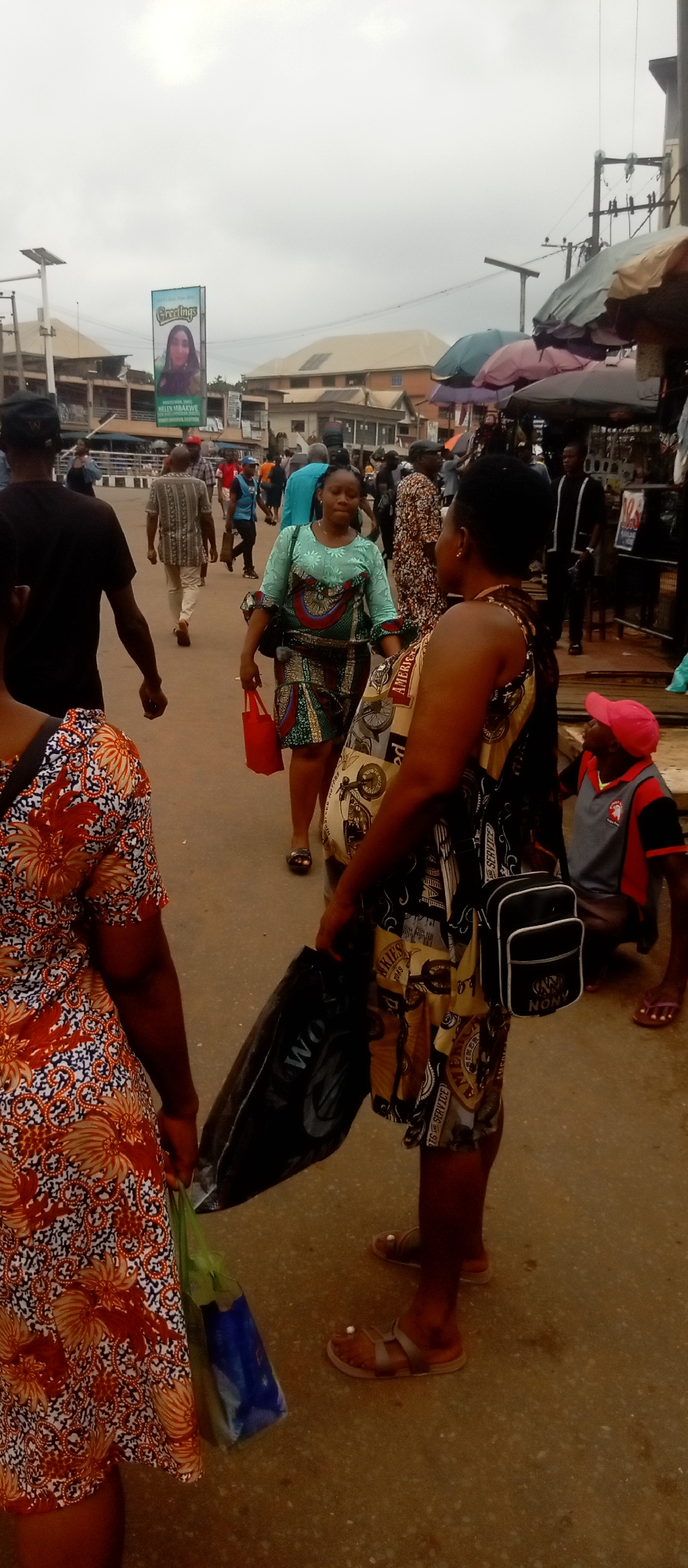 The image depicts a street scene in a crowded urban environment, likely a market or commercial district. The setting appears to be in Nigeria, Africa, given the clothing styles and the presence of a campaign poster with a Nigerian name and greeting.
Several individuals are visible: a woman in the foreground wearing a dress with a motorcycle pattern and carrying a black bag and small purse; another woman to her left in a floral orange dress; a woman in a green and patterned dress walking away; and a man crouching with a red cap. Other people are scattered in the background.
The environment includes buildings, some with metal roofs, and utility poles. Umbrellas provide shade for vendors. The sky is overcast, suggesting a cloudy day.