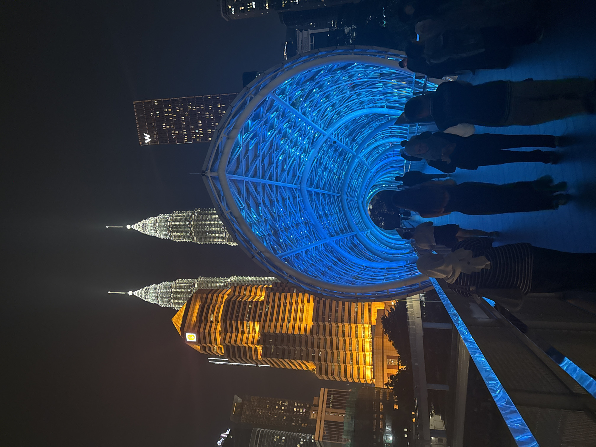 The image shows the Saloma Link pedestrian bridge in Kuala Lumpur, Malaysia, at night. The Petronas Twin Towers are visible in the background. A group of people is walking along the bridge, which is illuminated with blue lights. The bridge has a distinctive structural design.