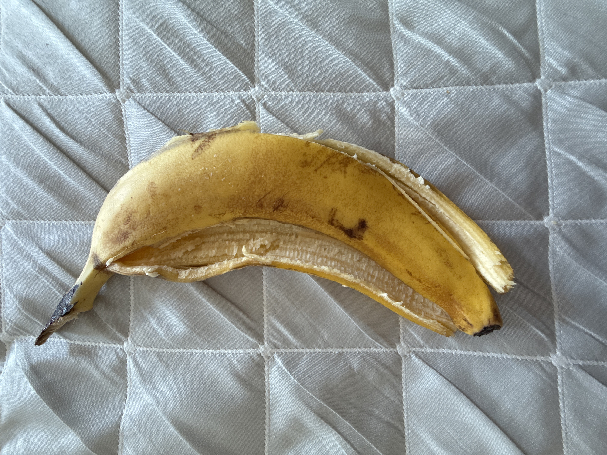 A discarded banana peel lies on a quilted, white fabric surface. The banana peel is yellow with brown spots and a dark, bruised tip. The peel is partially open, revealing the inside of the peel. No people, subjects, actions, or interactions are visible. The environment is limited to the immediate surroundings of the banana peel and the fabric. The location of the image is not in Izhevsk, Russia.