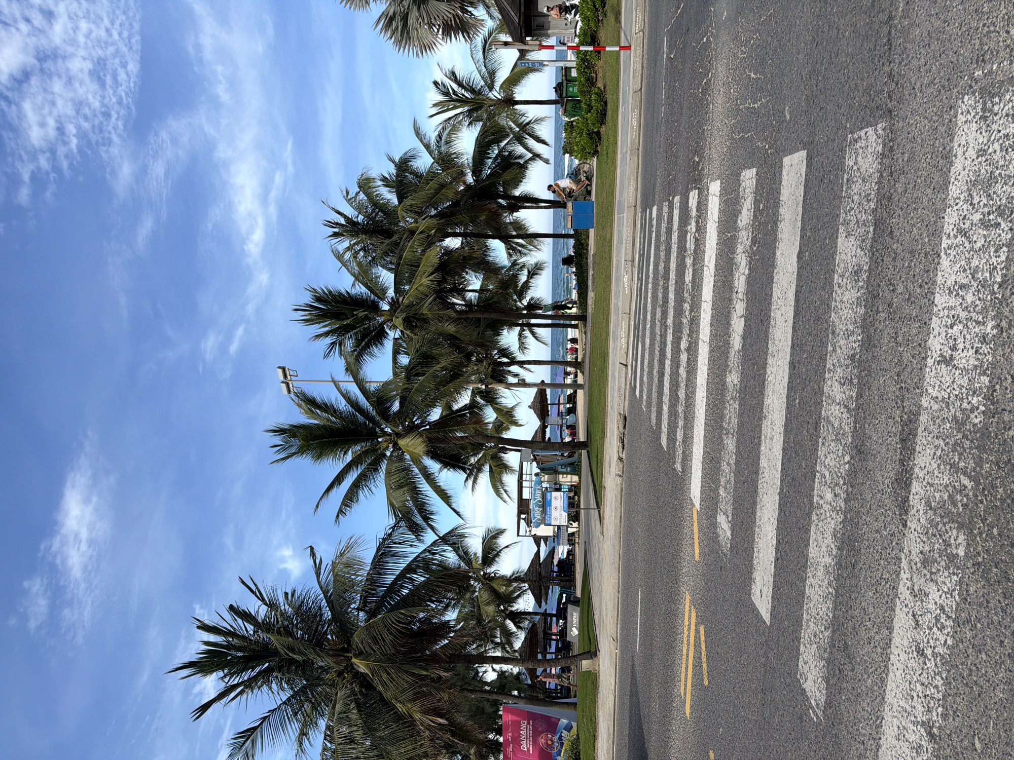 The image depicts a street scene in Đà Nẵng, Vietnam, adjacent to a beach. The view is oriented with the road running horizontally across the frame. A pedestrian crosswalk is prominently visible on the road surface, with white stripes contrasting against the asphalt. To the left of the road, palm trees line the beachside, partially obscuring the view of the beach and ocean. Under the trees, there are hints of beach umbrellas and people enjoying the beach. A sign, possibly advertising "Surf Shop," is also visible along the beachside. The sky is blue with scattered clouds, providing a backdrop to the palm trees. The road surface shows wear and tear, with yellow lines indicating traffic lanes.