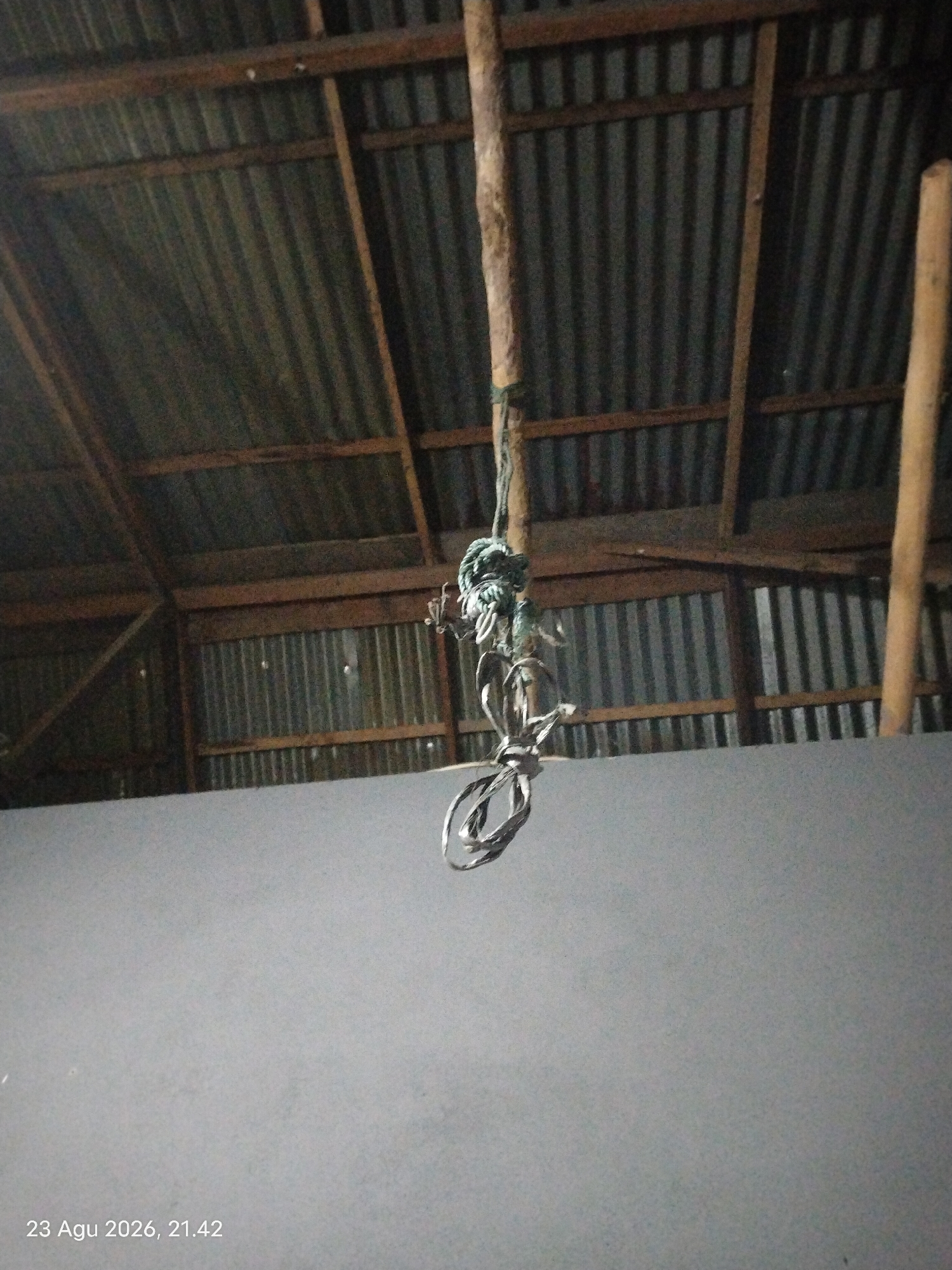 The image shows the interior of a structure with a corrugated metal roof and wooden beams. A thick rope is hanging from one of the beams, coiled and knotted. The rope appears to be made of natural fibers, possibly sisal or jute, and is greenish-blue in color. It is suspended over a grey, solid surface, possibly a wall or a platform. The background is dark and indistinct, with more corrugated metal visible. There are no people present in the image. The timestamp at the bottom of the image indicates it was taken on August 23, 2026, at 21:42. The location is identified as Singkawang, Indonesia.
