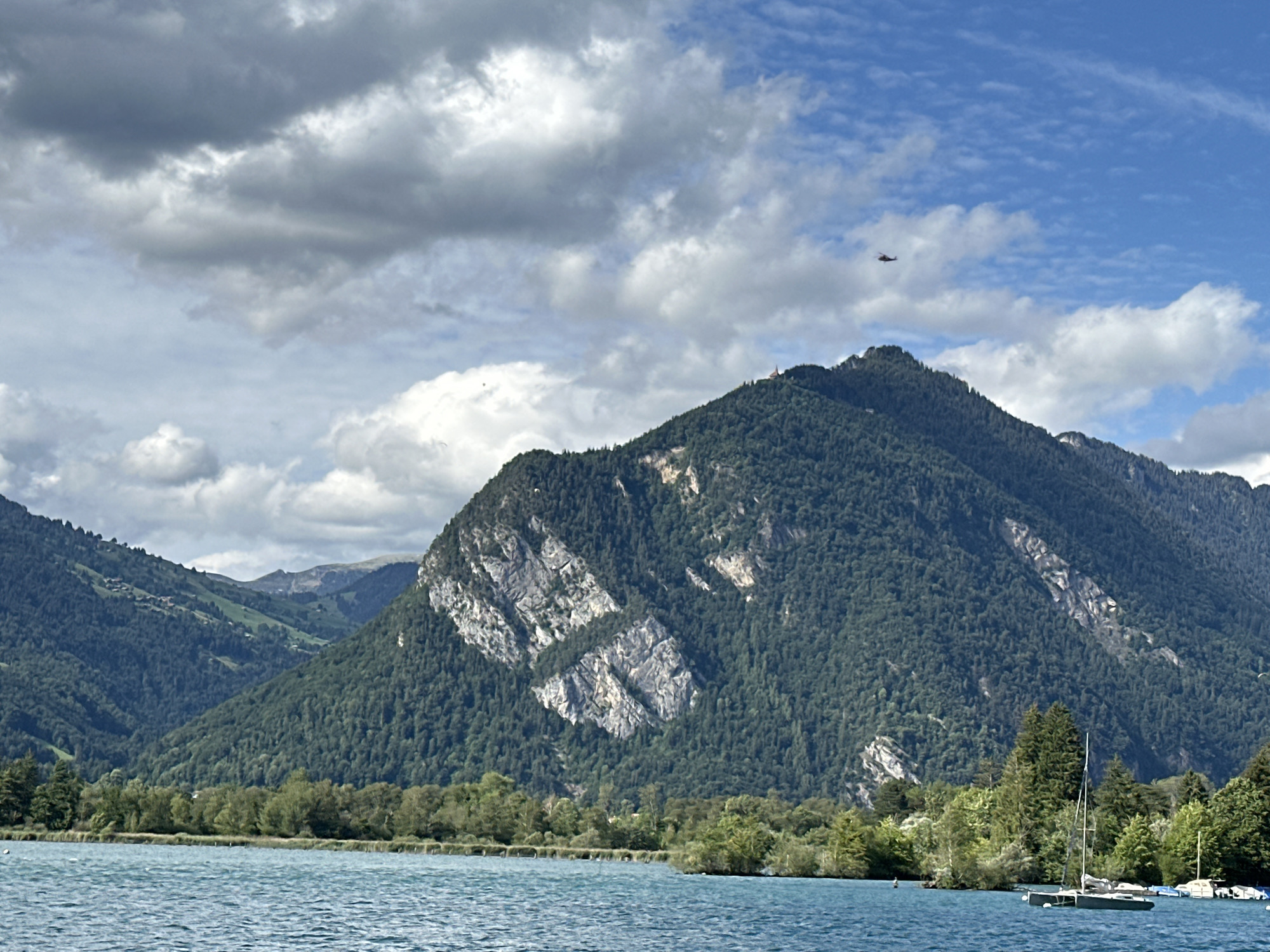 A wide, slightly low-angle shot captures a panoramic view of Lake Thun in the vicinity of Unterseen, Switzerland, on a partly cloudy day. The foreground is dominated by the turquoise water of the lake, with gentle ripples on its surface. A sailboat with a white sail, partially furled, is moored in the right mid-ground. Behind the lake, a dense line of trees forms the shoreline, transitioning into the steep, tree-covered slopes of the mountains. The dominant mountain in the center of the frame features a prominent, light-colored rocky outcropping. In the distance, a smaller helicopter is visible in the bright blue sky, a few white clouds also present. The overall scene is natural and serene.
