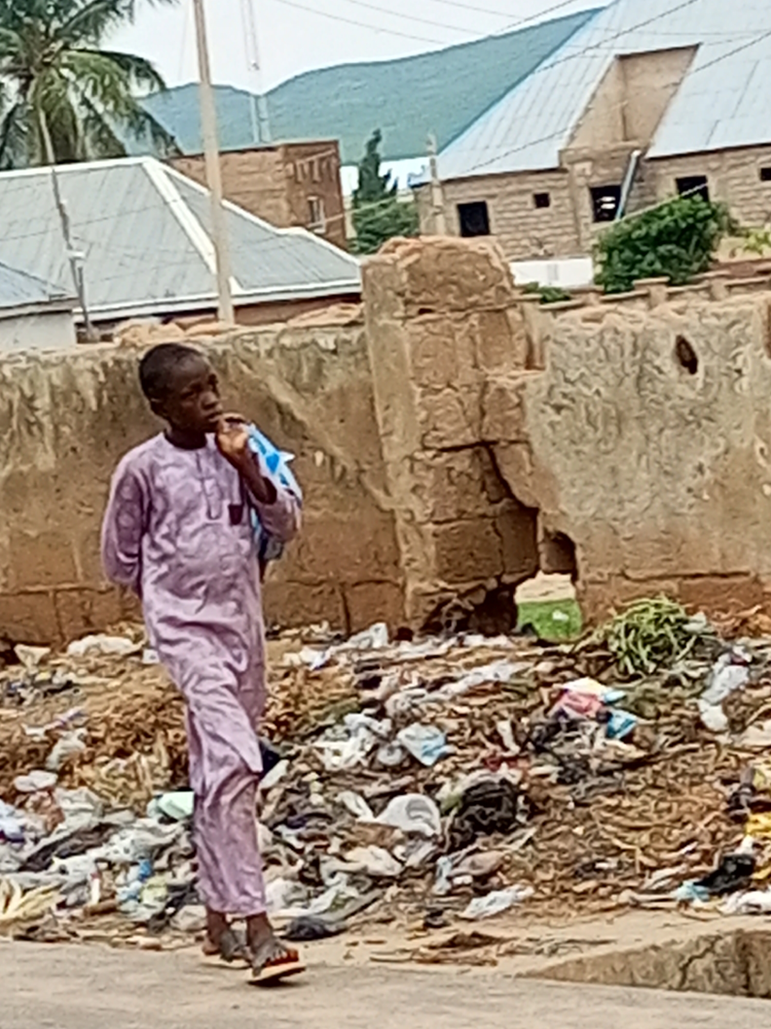 A young boy, appearing to be of African descent, walks along a dirt path in Arawa, Nigeria. He is wearing a long, light purple robe with intricate patterns and sandals. He is carrying a blue and white plastic bag over his left shoulder and appears to be holding something to his mouth with his right hand. The boy is walking past a large pile of assorted garbage, including plastic bags, fabric scraps, and other debris. Behind the garbage pile is a crumbling, rough-hewn wall made of earth or mud bricks. Further in the background, there are buildings with corrugated metal roofs and some greenery, including a palm tree on the left. The sky is overcast.