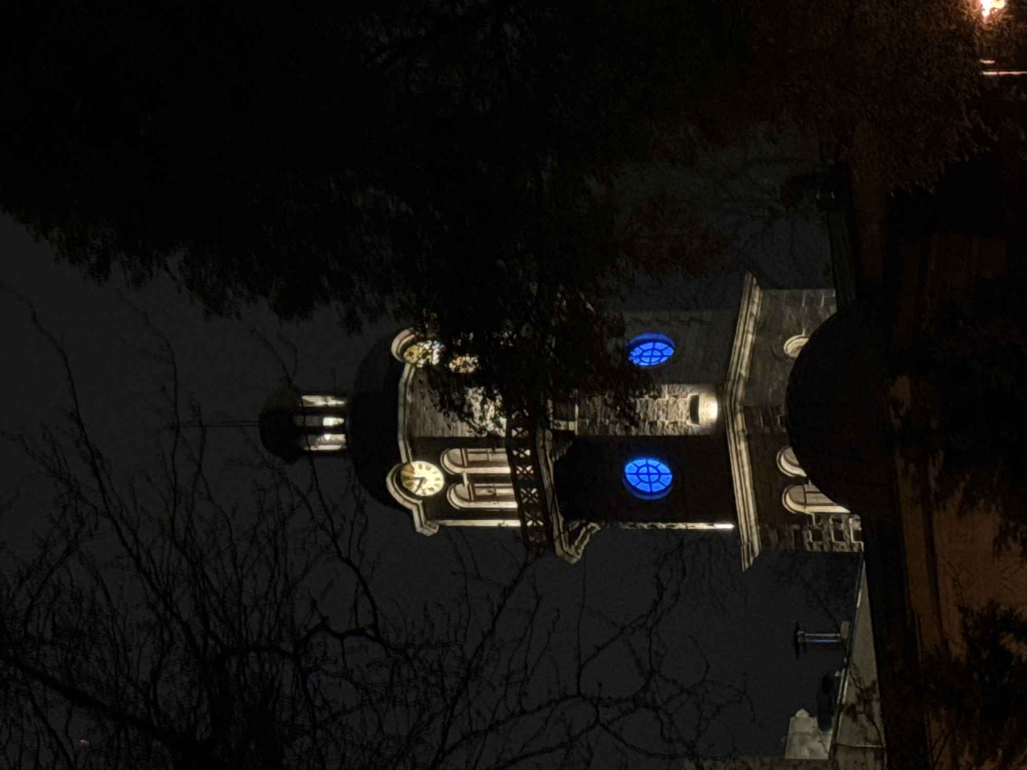 At night, a low-angle shot shows a building, possibly a church, located in Varna, Bulgaria. The building features a tower with a clock face and two blue circular windows. The lower part of the structure is obscured, but elements such as walls and possibly a roofline are visible. The architectural style is indicative of historical European construction. Bare tree branches are prominent in the foreground and on the sides. The lighting is dim and artificial, accentuating the building’s key features while leaving other areas in shadow. A nearby structure is seen on the right with illumination at the top.