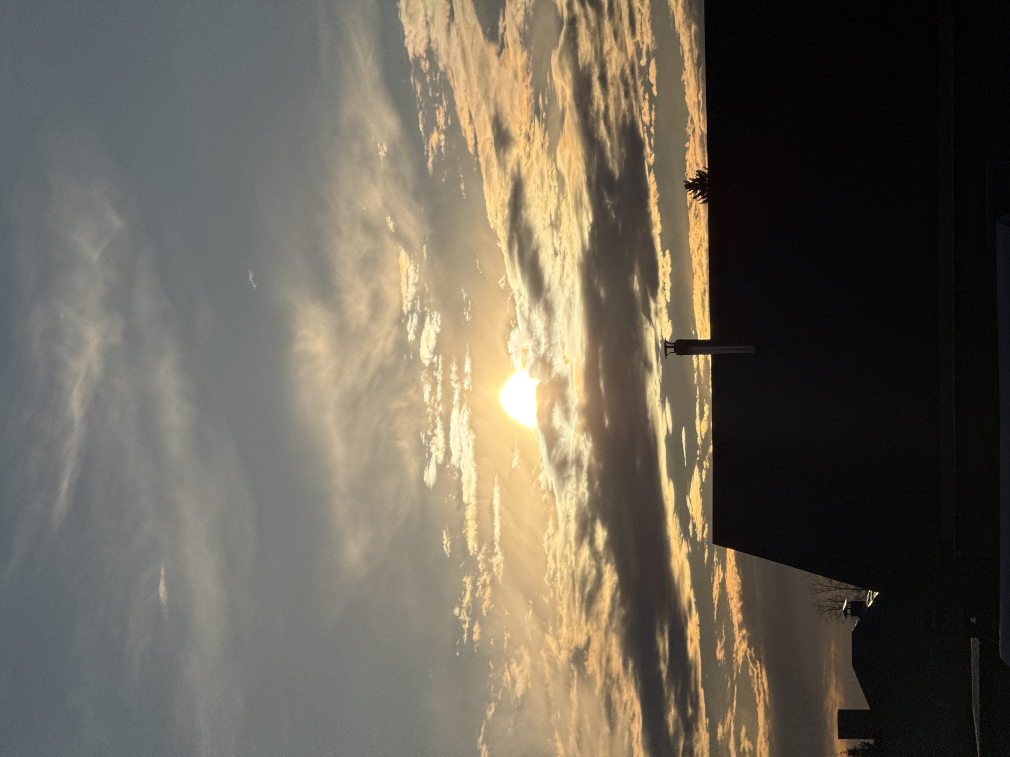 The image shows a sunset with clouds in the sky. In the foreground, there is a dark silhouette of a building and what appears to be a utility pole or other structure extending upwards. It's impossible to determine the specific location in Chaikovsky, Russia from the given image. There are no people or other subjects visible beyond the sky and the vague silhouettes of the manmade structures.