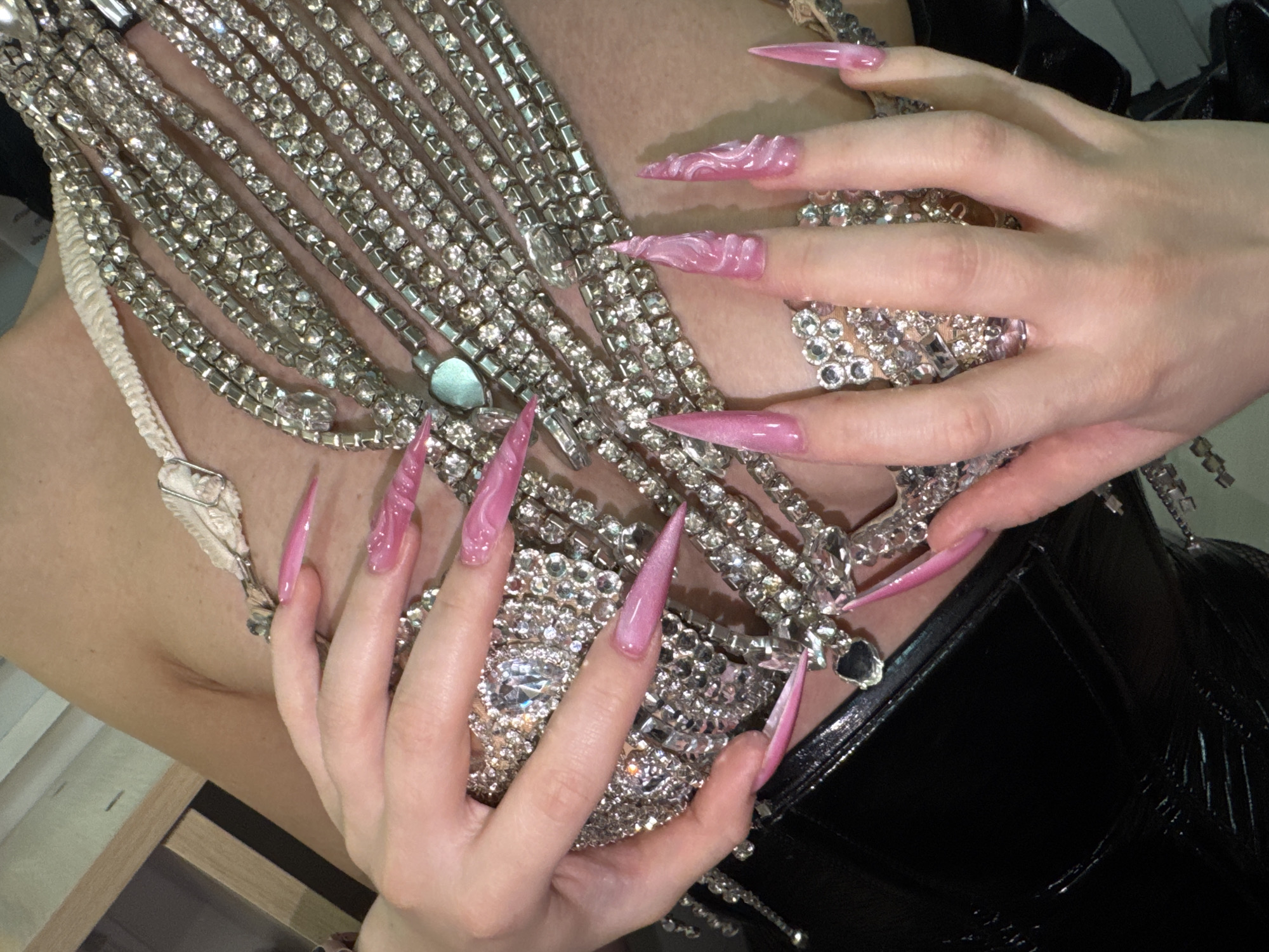 The image shows a close-up view of a person's chest and hands adorned with elaborate jewelry and striking pink nails. The jewelry consists of multiple strands of sparkling rhinestones that form a decorative bodice or necklace. One hand is positioned over the chest, touching the jewelry, while the other hand is partially visible, also interacting with the jewelry. The nails are long, pointed, and painted in a vibrant pink with a swirling, marbled design. The person is wearing a black, glossy garment, possibly a corset or top. The background is out of focus, but appears to be an indoor setting with a light-colored wall and some wooden furniture. The image does not contain any recognizable landmarks or features that would suggest a specific location within the city of Kazan, Russia.