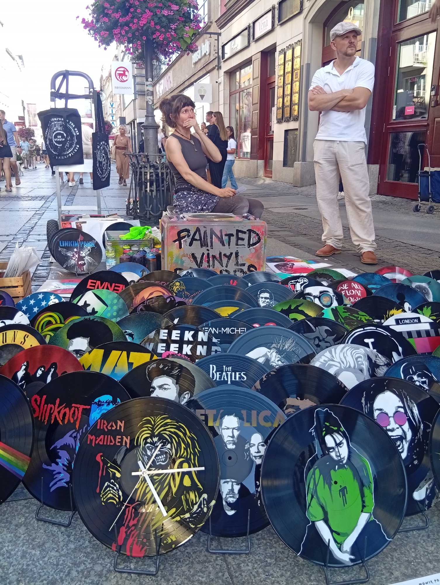 The image depicts a street market scene in Wrocław, Poland, likely on a cobblestone street given the paving. In the foreground, a large collection of painted vinyl records is displayed on the ground and on stands. These records feature various popular music artists and bands such as Iron Maiden, Slipknot, The Beatles, and Billie Eilish, along with iconic imagery like the American flag and a rainbow.
A woman is seated on a low stool or box, which is also painted with the words "PAINTED VINYL". She is looking to the left with her hand near her mouth, appearing to be resting or interacting with someone out of frame. She is wearing a dark top and a patterned skirt.
Behind the records and the woman, a man in a light-colored flat cap, white polo shirt, and beige trousers stands with his arms crossed, looking to his left. His stance suggests he might be a vendor or a passerby observing the scene.
In the background, the street continues with more people walking, suggesting a public thoroughfare. Storefronts with awnings and signage are visible on the buildings lining the street. A lamppost with hanging pink flowers stands near the woman. The overall atmosphere suggests a lively outdoor market.