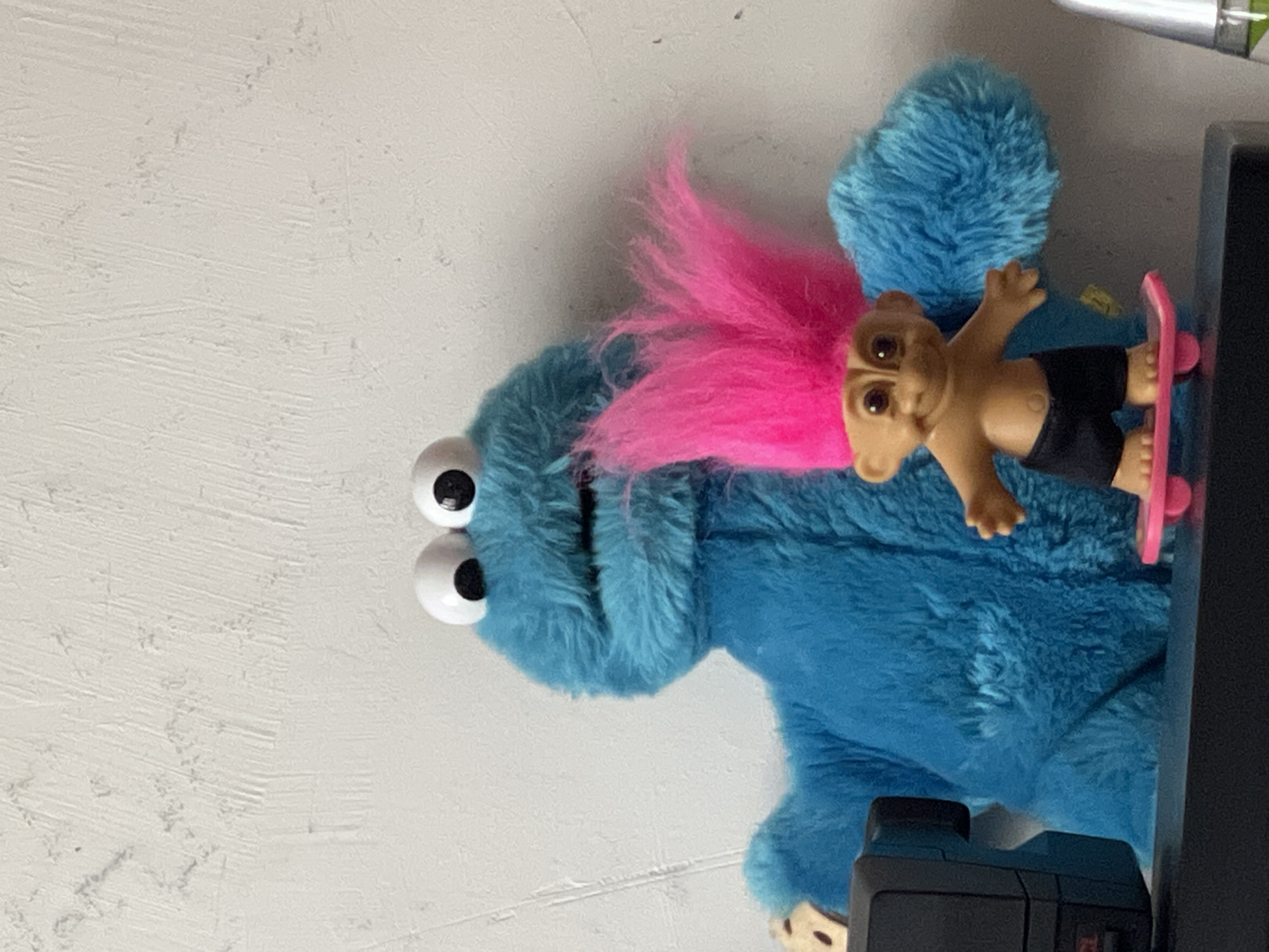 A blue Cookie Monster plush toy sits against a white wall. On top of the Cookie Monster is a troll doll with pink hair, standing on a pink skateboard. The troll doll is wearing black shorts. The location is not identifiable in the image and the image could not have been taken in Moscow, Russia based on the objects in the image.