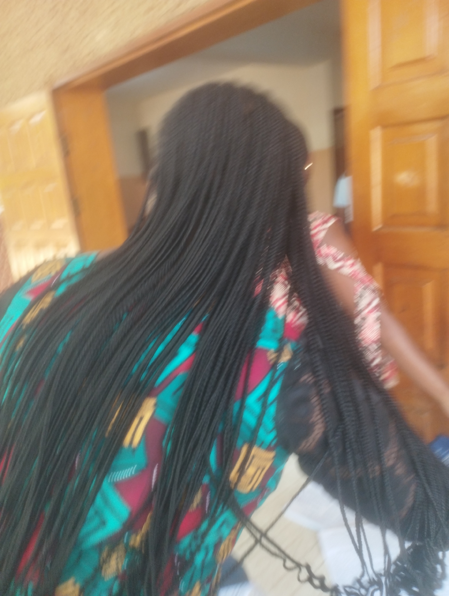 In the city of Jos, Nigeria, the image captures the back of a person wearing a vibrantly patterned shirt with teal, red, and yellow geometric designs. Their long, braided black hair falls down their back. The person is standing near a wooden door or partition, with another person's arm, also adorned with a similar patterned sleeve, visible in the background. The environment suggests an indoor setting with wooden elements, possibly a home or public building.