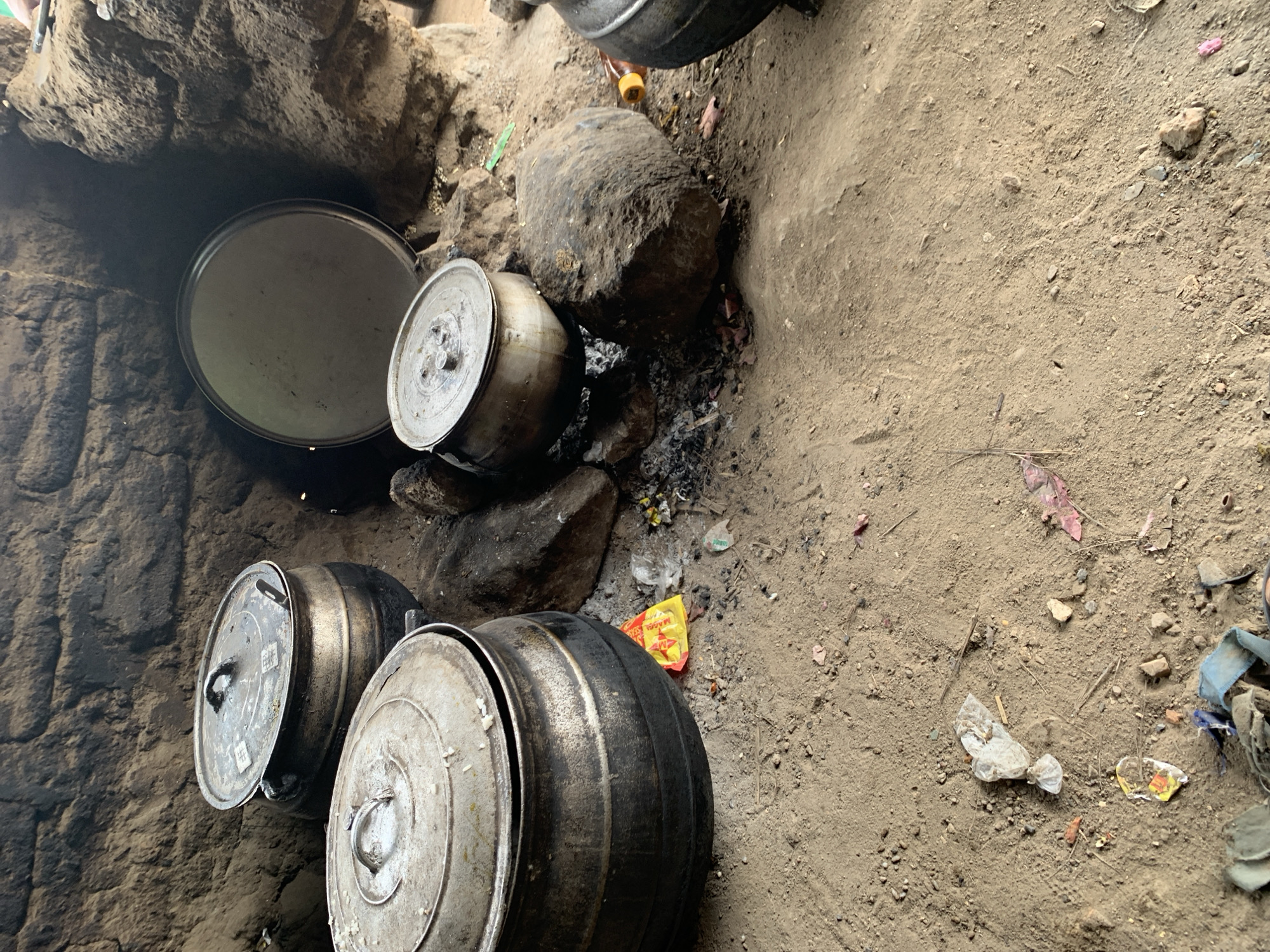 The image depicts an outdoor cooking area in what appears to be the city of Dull A, Nigeria. The scene is characterized by a rustic, earthy environment with a dirt floor and a backdrop of rough, textured walls, possibly made of mud or clay. Several large, dark metal pots are present, some with lids, indicating a cooking process is underway or has recently concluded.
In the foreground, a large pot with a lid is prominently positioned. To its left, another similar pot is visible, also with its lid. In the background, a large, flat, circular metal object, possibly a tray or a lid, rests against a rocky formation. Adjacent to the cooking pots, there are several large rocks, which may be used to support the cooking vessels or as part of the hearth. Scattered on the ground around the pots are remnants of food, ash, and debris, including small pieces of plastic and paper packaging, suggesting a meal preparation area.
The overall impression is one of a traditional, perhaps communal, cooking setup in an open-air environment. The lighting suggests it is daytime, with some shadows indicating the direction of light. There are no people visible in the image.