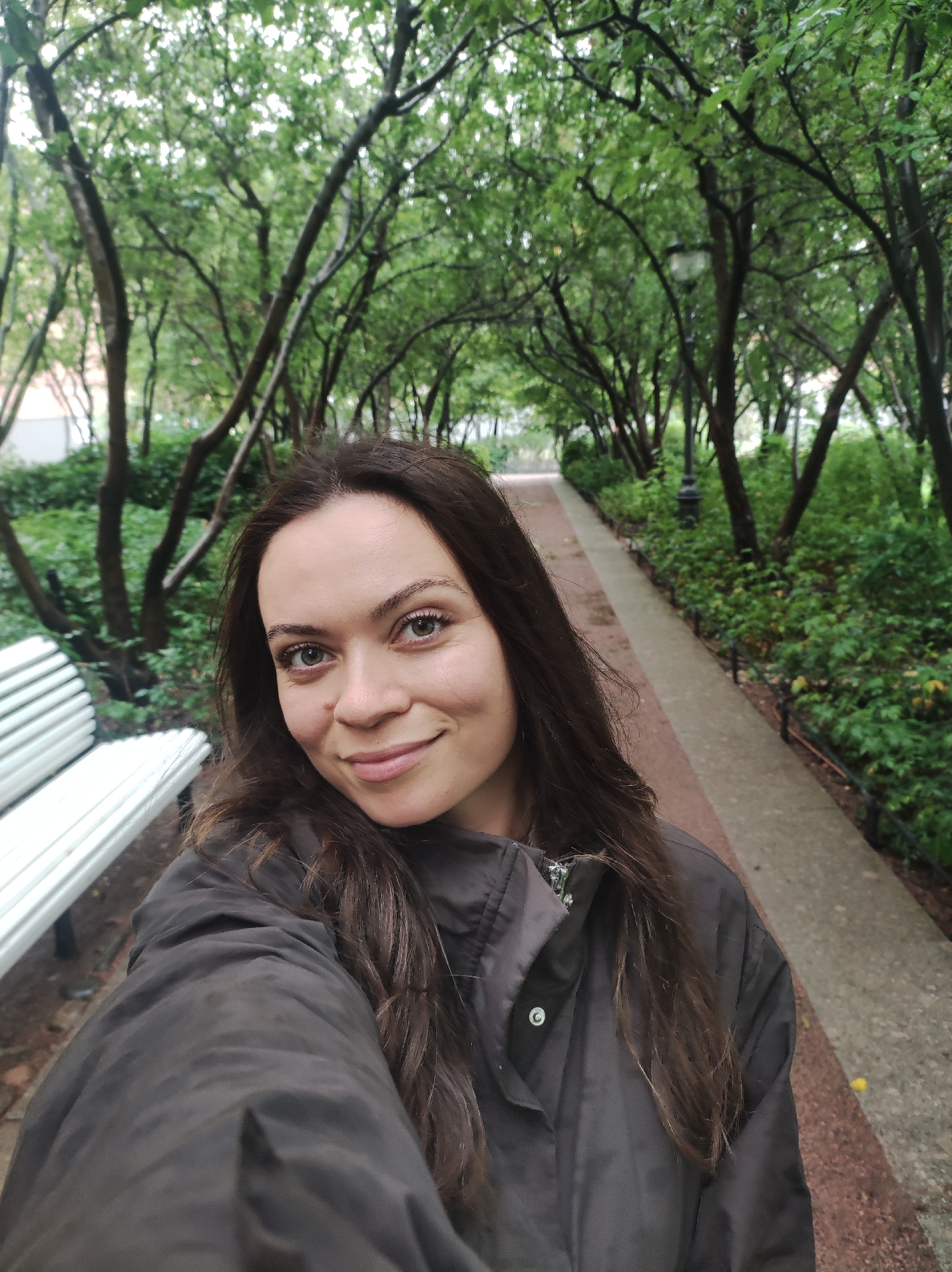 A medium shot photograph shows a young Caucasian woman with long, dark brown hair and light green eyes. She is looking directly at the camera with a slight smile and appears to be taking a selfie. She is wearing a dark-colored puffer jacket.
To her left, a portion of a white park bench with a slatted seat is visible.
The background depicts a tree-lined path with a paved walkway. The trees are dense with green foliage, suggesting a park or garden setting. A lamppost is visible further down the path. The overall atmosphere is overcast, and the ground appears damp, indicating recent or ongoing rain.
The scene is consistent with an outdoor public park in Saint Petersburg, Russia, possibly during the spring or summer months.