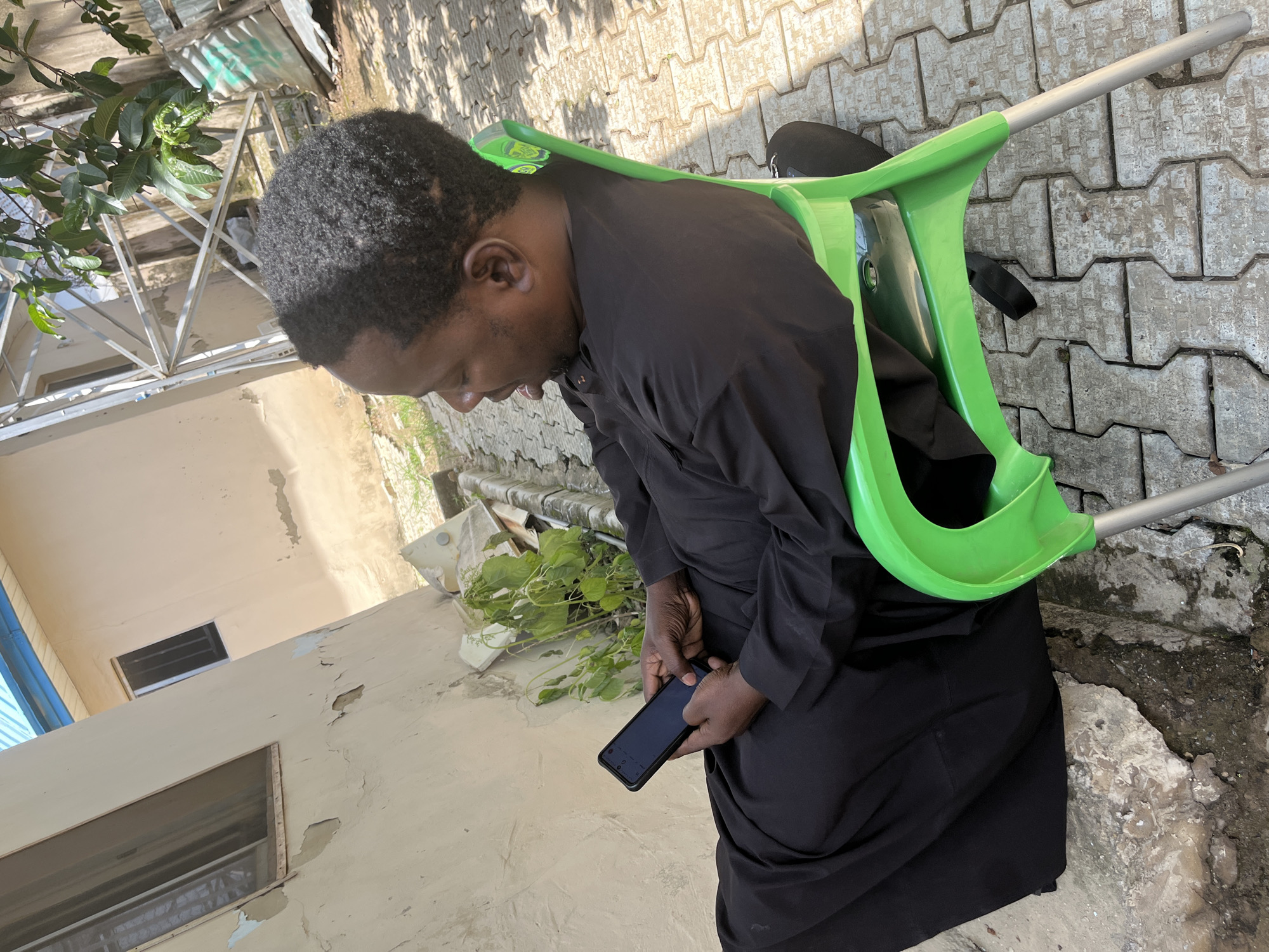 A man is seated on a bright green plastic chair in Galadimawa Village, Nigeria. He is wearing a black traditional outfit and is looking down at a black smartphone in his hands. The man's hair is short and dark. He is sitting on a paved area made of interlocking concrete blocks. Behind him, a light-colored building with peeling paint and a window is visible. Overhead, there are branches of a tree with green leaves, and a metal structure can be seen. Sunlight casts shadows on the ground, indicating it is daytime.