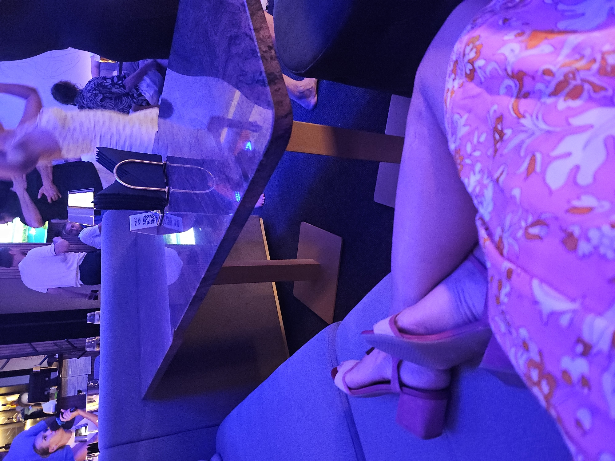 The scene is a lounge or bar, bathed in blue lighting. The focus is on a person seated on a grey-blue cushioned bench, their crossed legs prominently displayed. They wear a pink floral dress and reddish-brown block-heeled sandals. A marble-topped table with menus sits in front of them, the table reflecting the lighting in a distorted manner. Other people are present in the background, including one individual holding a microphone, suggesting possible karaoke.