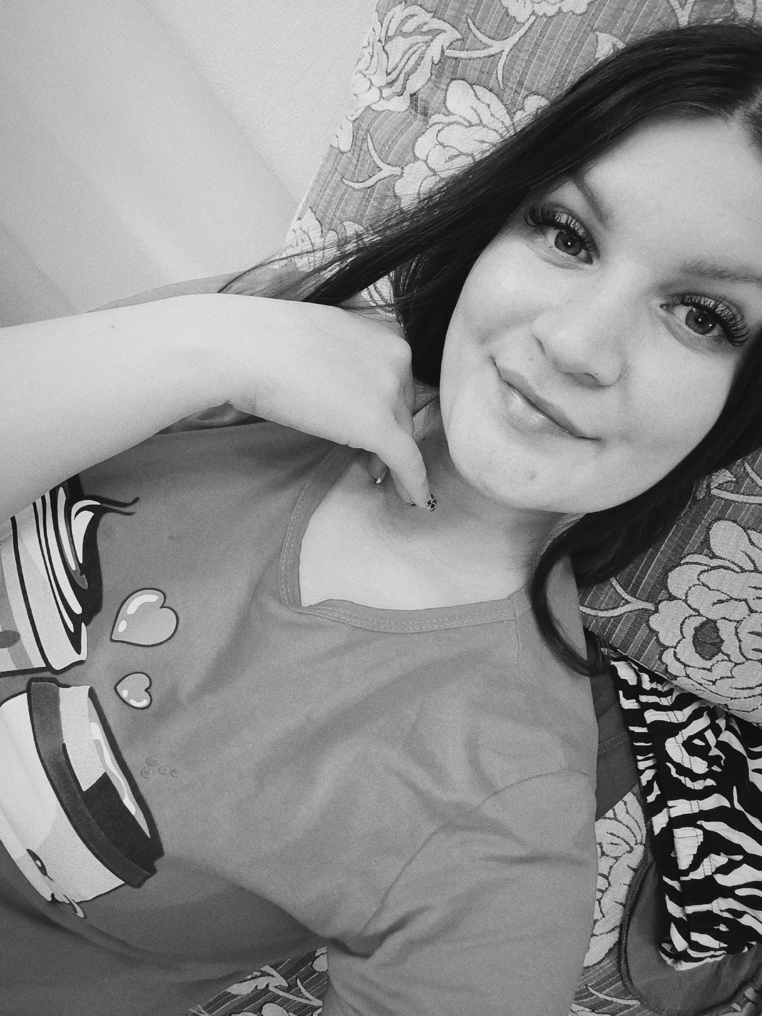 A young woman is depicted in a black-and-white photograph, lying on a patterned surface, possibly a bed or sofa. She has dark hair, eyelashes, and is wearing a t-shirt with a graphic design that includes heart shapes. Her left hand is raised and positioned near her neck. The patterned surface beneath her features a floral design. A separate piece of material with a zebra print is also visible. The photograph appears to be taken in an indoor setting. There is no information to indicate the location of the city of Tobolsk, Russia.
